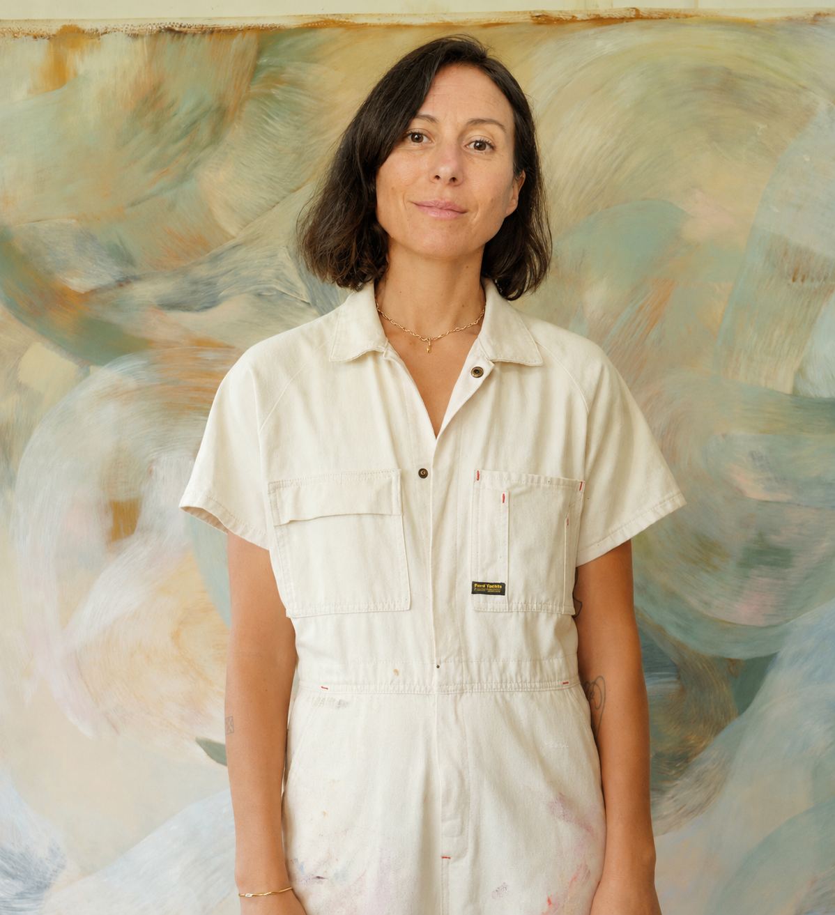 Une femme avec des cheveux bruns courts, portant une combinaison beige, posant devant une œuvre d'art abstraite aux couleurs pastel. Emilei Vincent, Yoga Aix-en-provence.