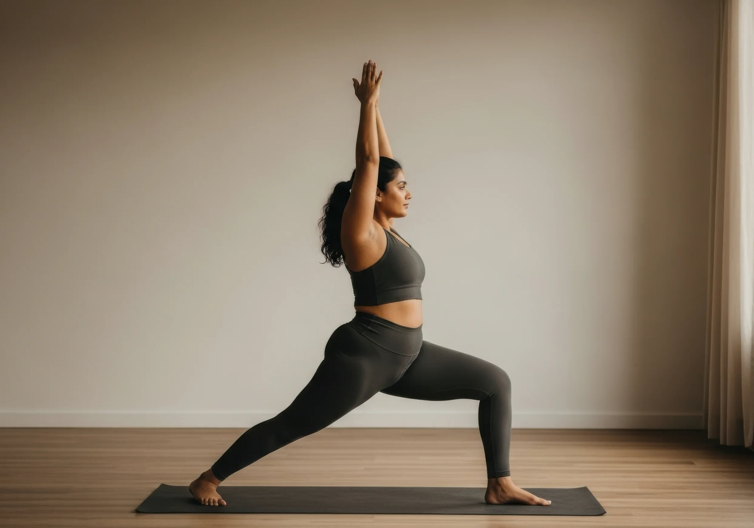 Femme pratiquant le yoga en position de guerrier avec une jambe pliée à 90 degrés et les bras levés, dans une pièce lumineuse avec un mur beige et un sol en bois.