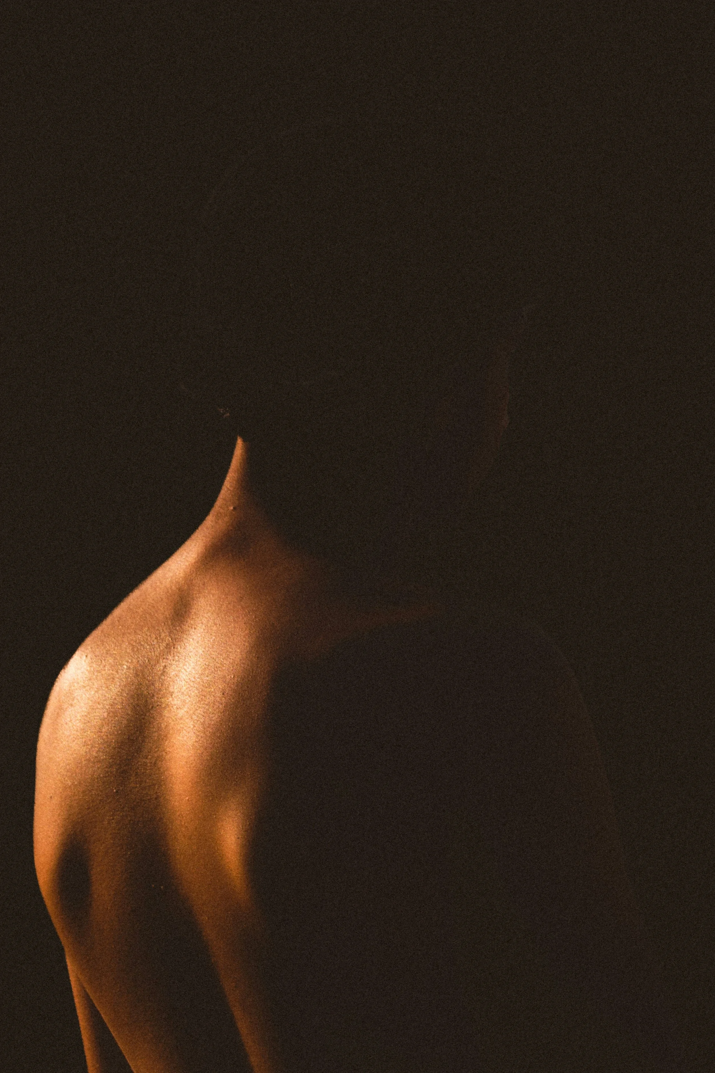 Une personne nue vue de dos, avec uniquement le haut du corps visible, dans une lumière douce et sombre.