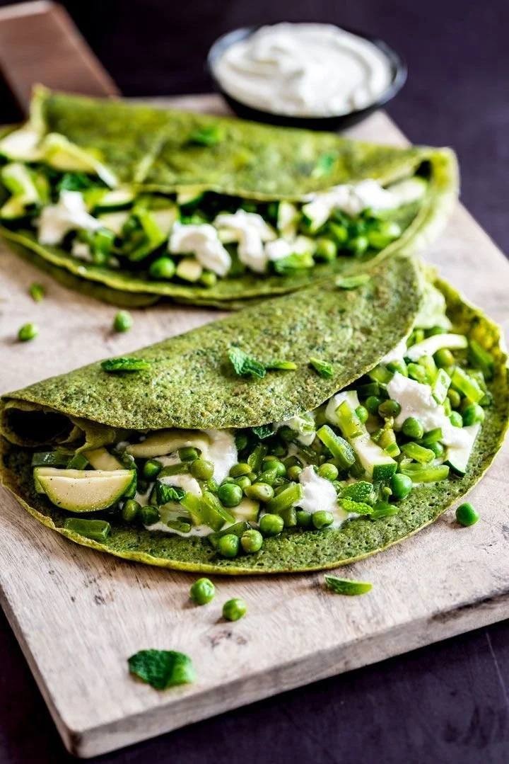 Deux tacos végétariens avec tortilla verte, garnis de pois verts, concombre, oignon et sauce blanche sur une planche en bois.