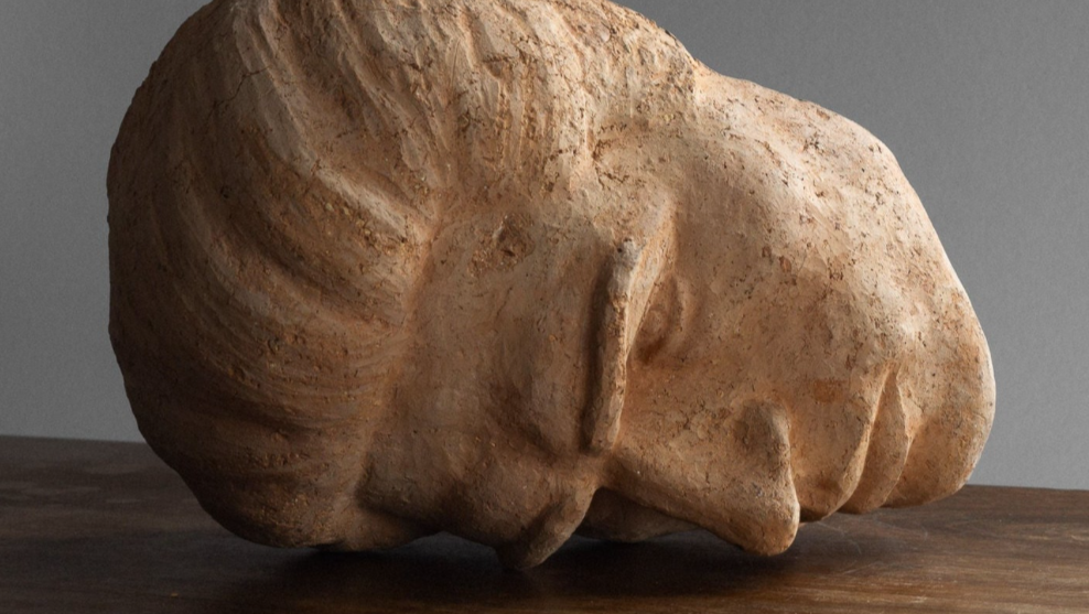 Une sculpture en pierre représentant une tête humaine. La tête est inclinée vers le bas, avec des détails stylisés du visage.