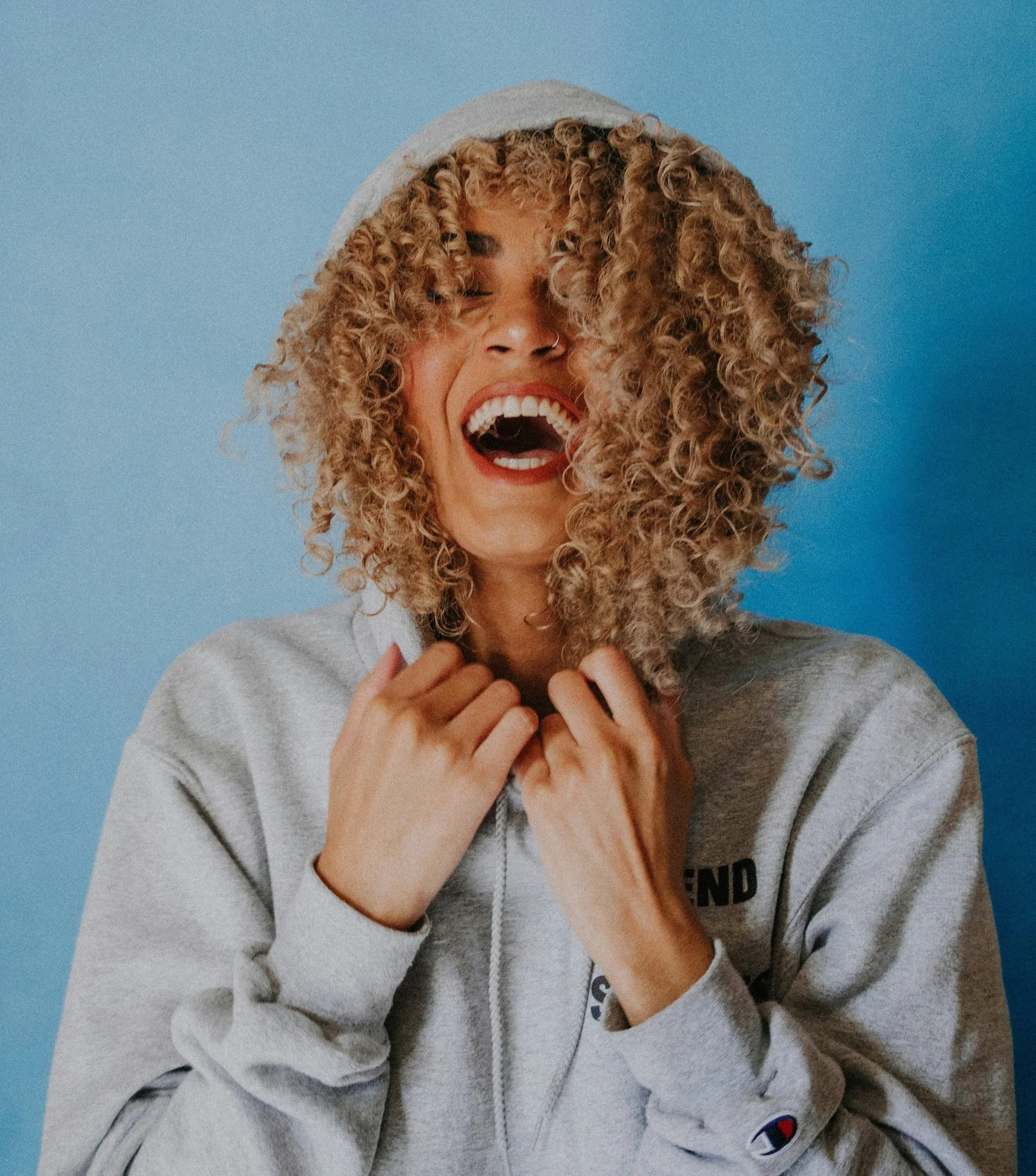 Une femme souriante portant une casquette gris clair et une veste gris clair, avec des cheveux bouclés, contre un fond bleu.
