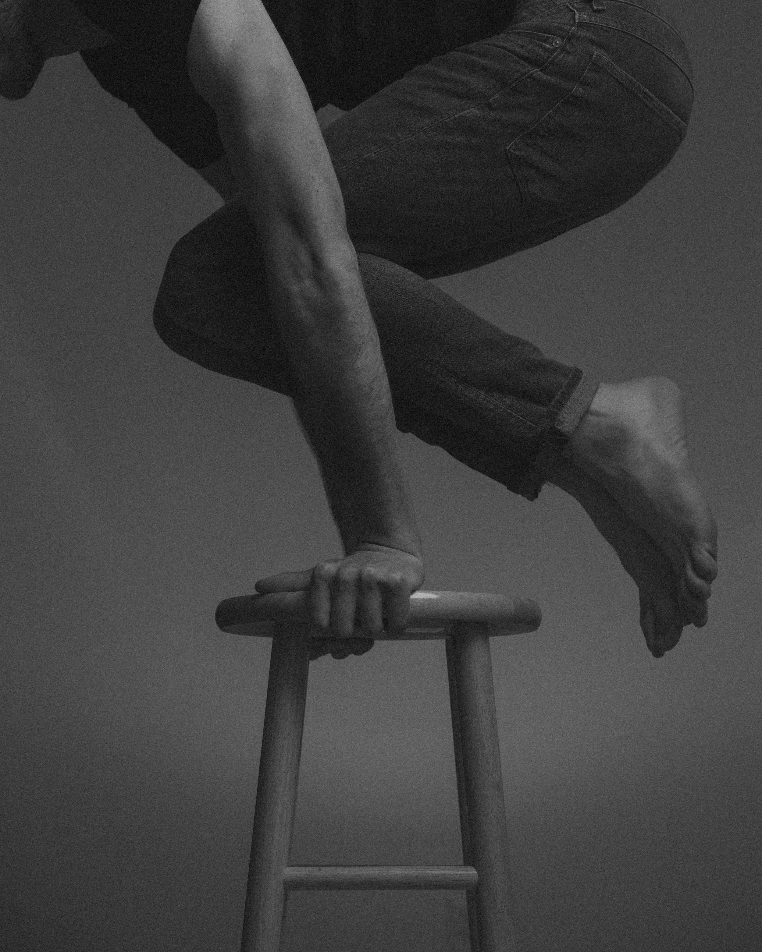Photo en noir et blanc d'une personne fragile en train d'équilibrer son corps sur une chaise en bois, utilisant sa main pour se soutenir.