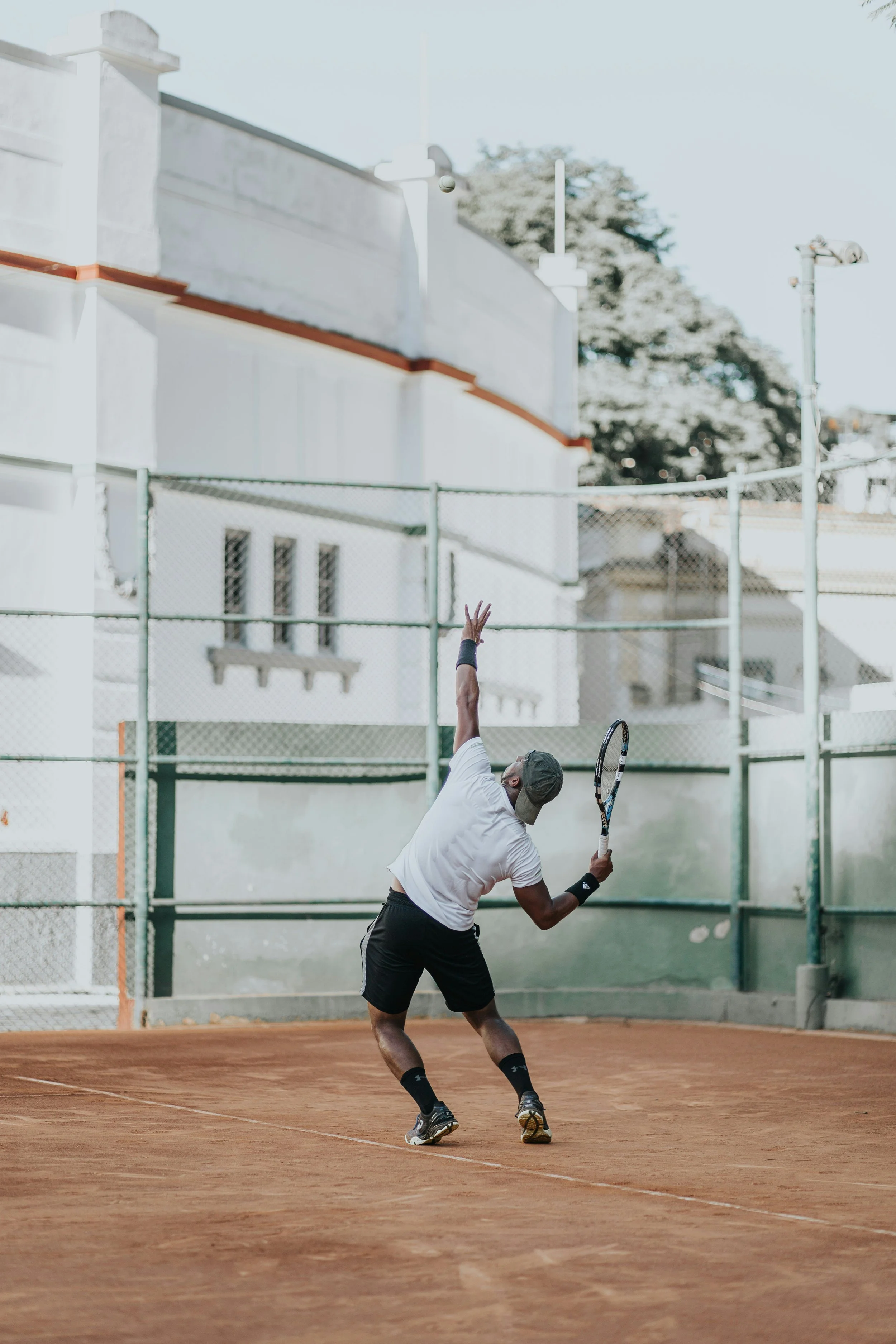 Un homme joue au tennis sur un court en terre battue, tenniskit blanc, tenant une raquette, en pleine action, fond avec clôture, bâtiments, et arbres.
