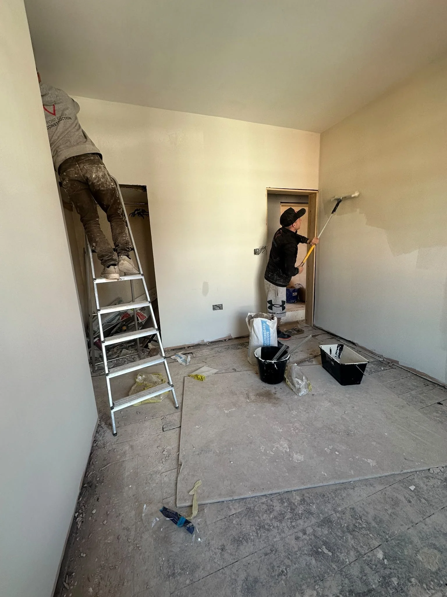 Two workers painting a wall in a room under renovation. One is standing on a ladder and the other is using a roller to apply paint.