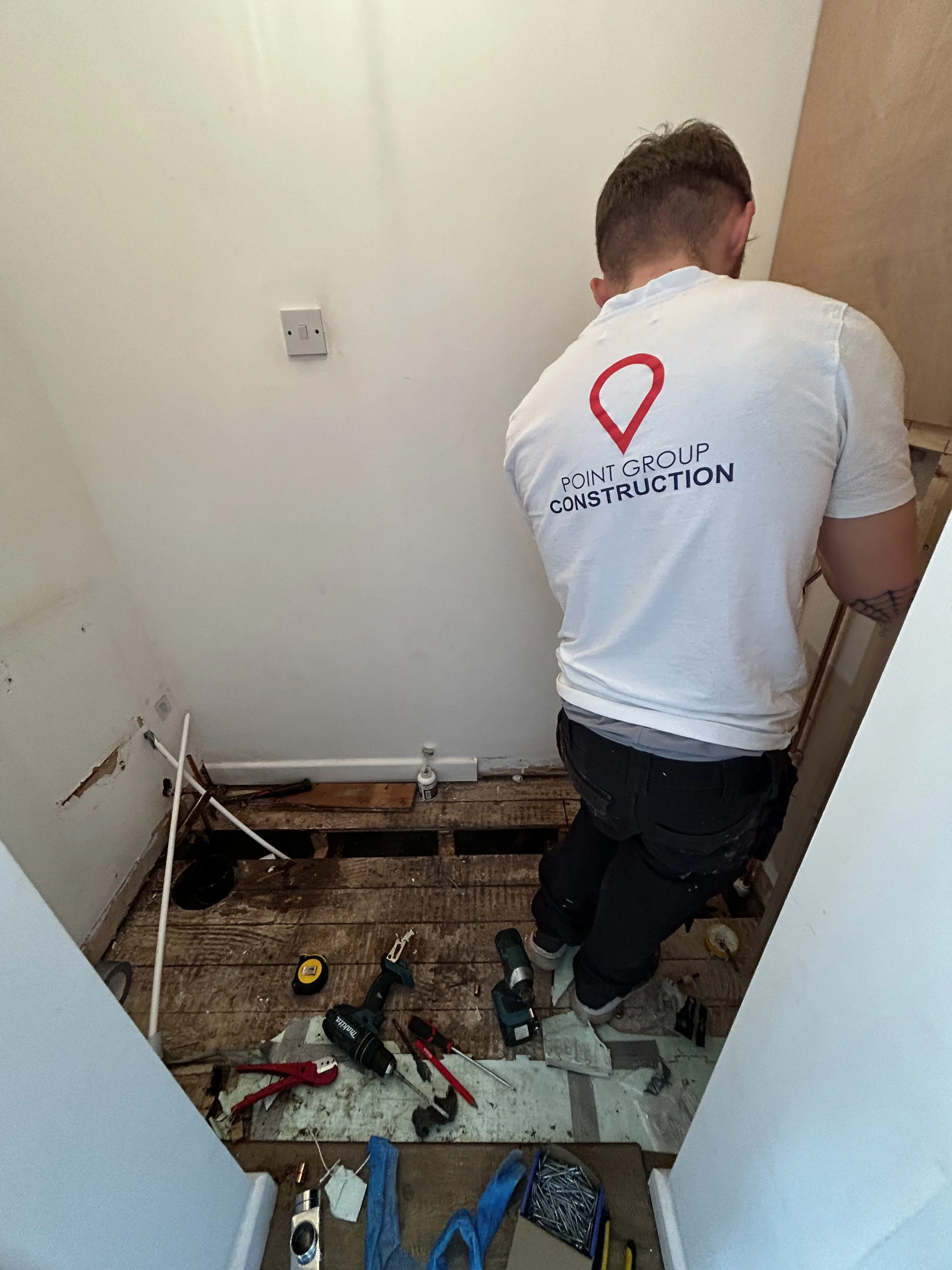 A person dressed in work clothes with a logo on the back reading 'Point Group Construction' is working on a home improvement project. They are facing away from the camera, working in a small, unfinished space with construction tools and materials on the floor, inside a room with a white wall and some exposed pipes.