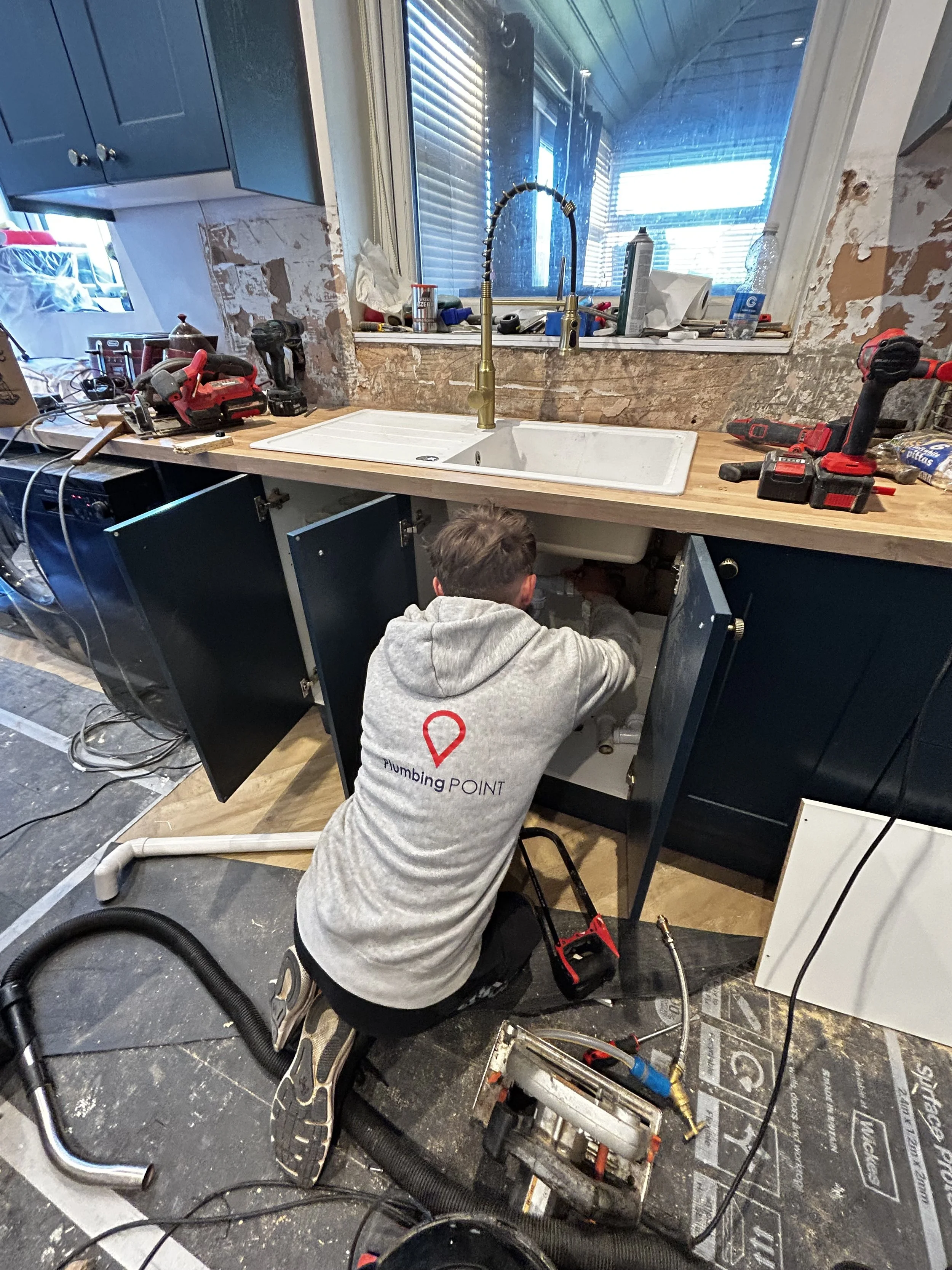 A plumber working under a kitchen sink with tools and equipment around, in a kitchen under renovation.