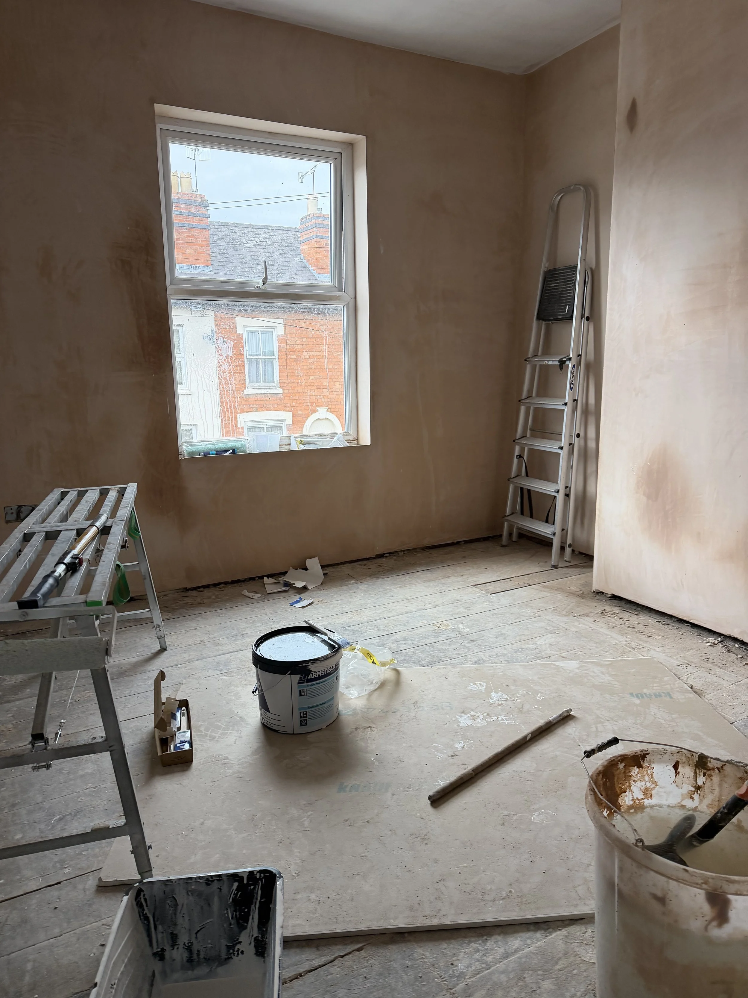 Empty room under renovation with a window showing neighboring brick buildings, ladder, paint buckets, and construction tools and supplies scattered on the unfinished wooden floor.