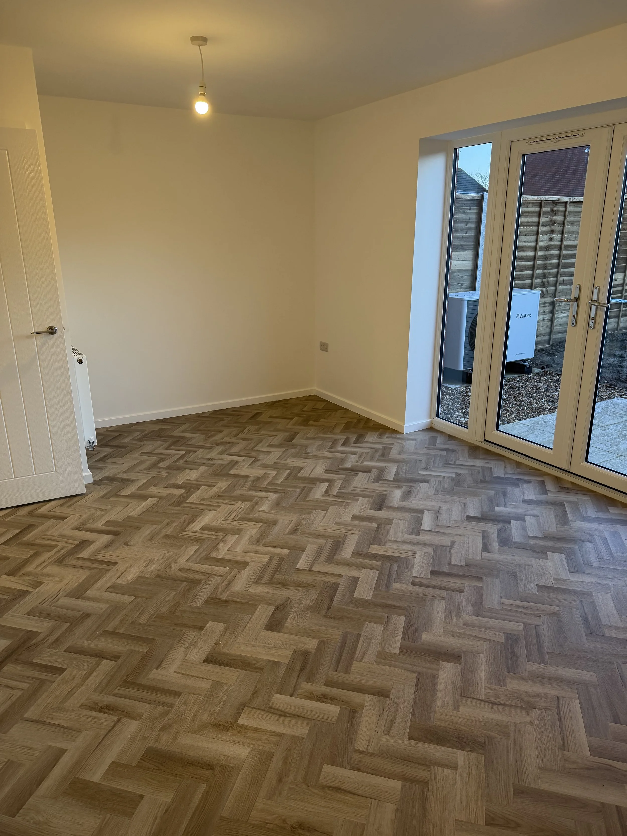 Empty room with wooden herringbone flooring, white walls, a large window with glass doors leading outside, and a ceiling light bulb.