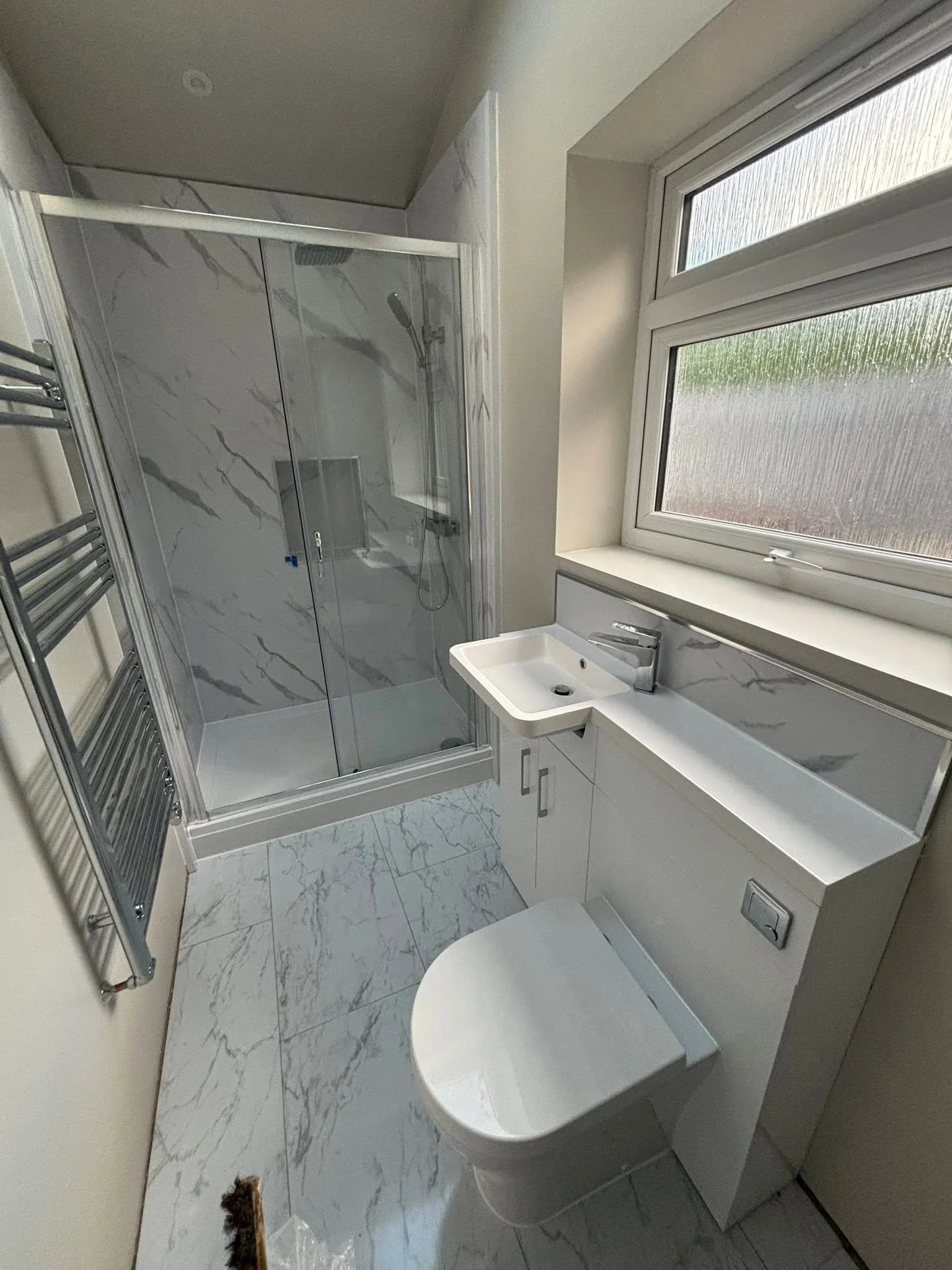 Bathroom with a shower, white sink, and toilet. Large window with textured glass, marble-patterned floor and shower walls, towel rack, and small decorative object near the toilet.