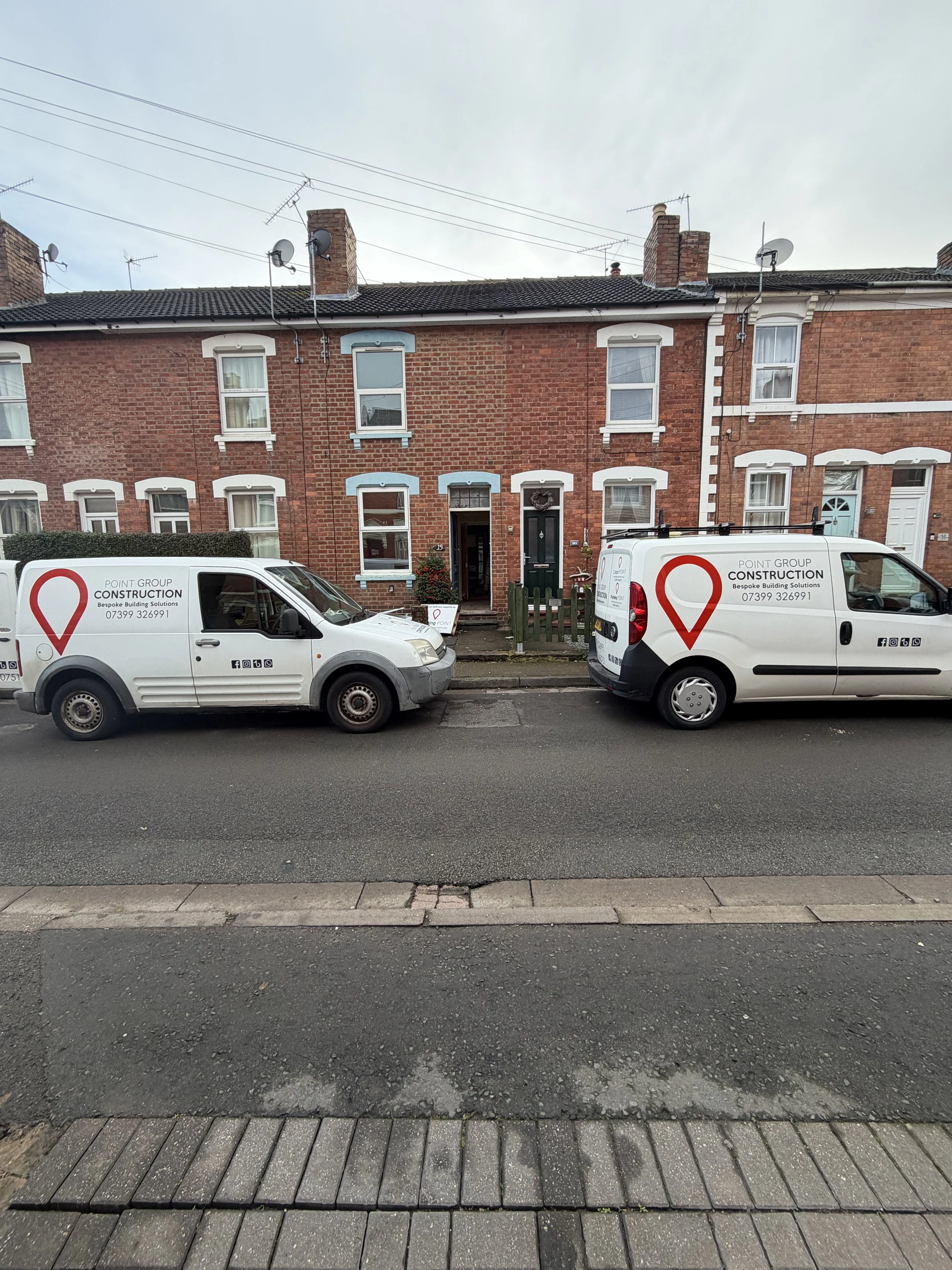 Two white company vans parked in front of a row of brick houses, with a grey sidewalk and a paved street in the foreground.