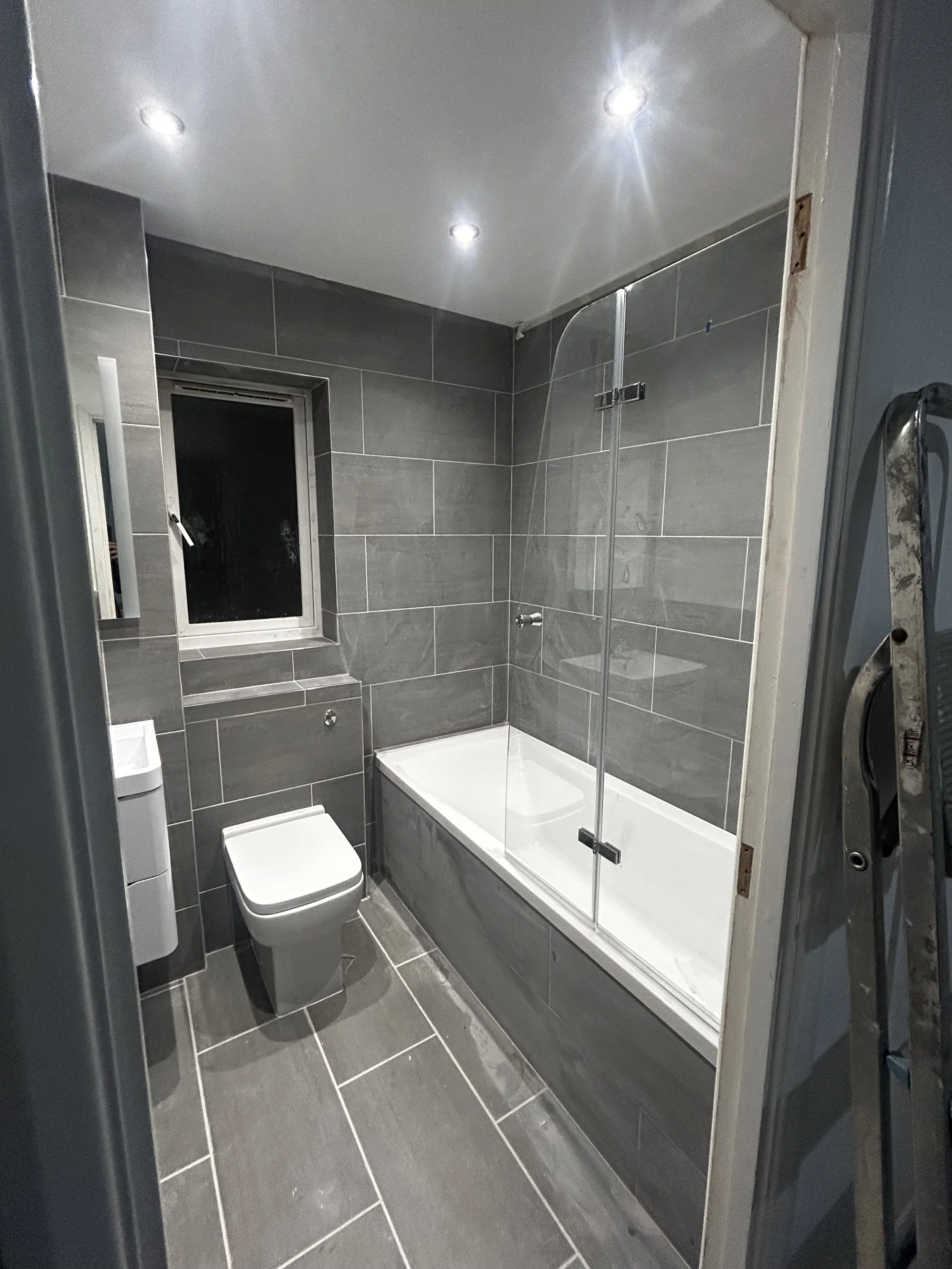 A modern bathroom with gray tiles on the walls and floor, a white bathtub, a gray toilet, a small window, and recessed ceiling lights.