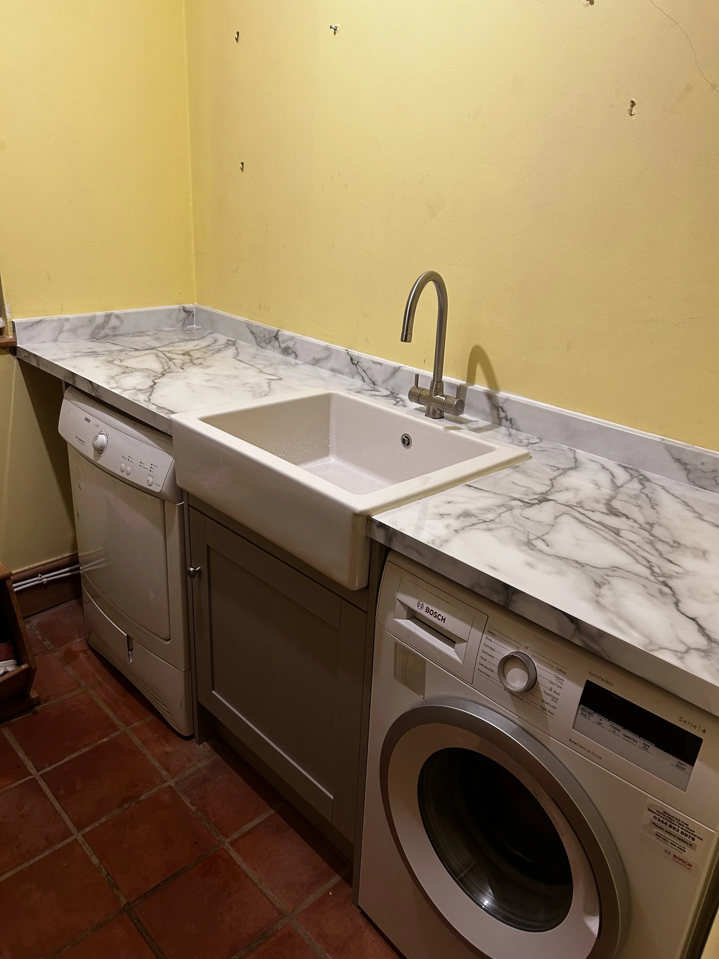 A laundry room with a yellow wall, marble countertop, white farmhouse sink, and a white washing machine below the countertop.