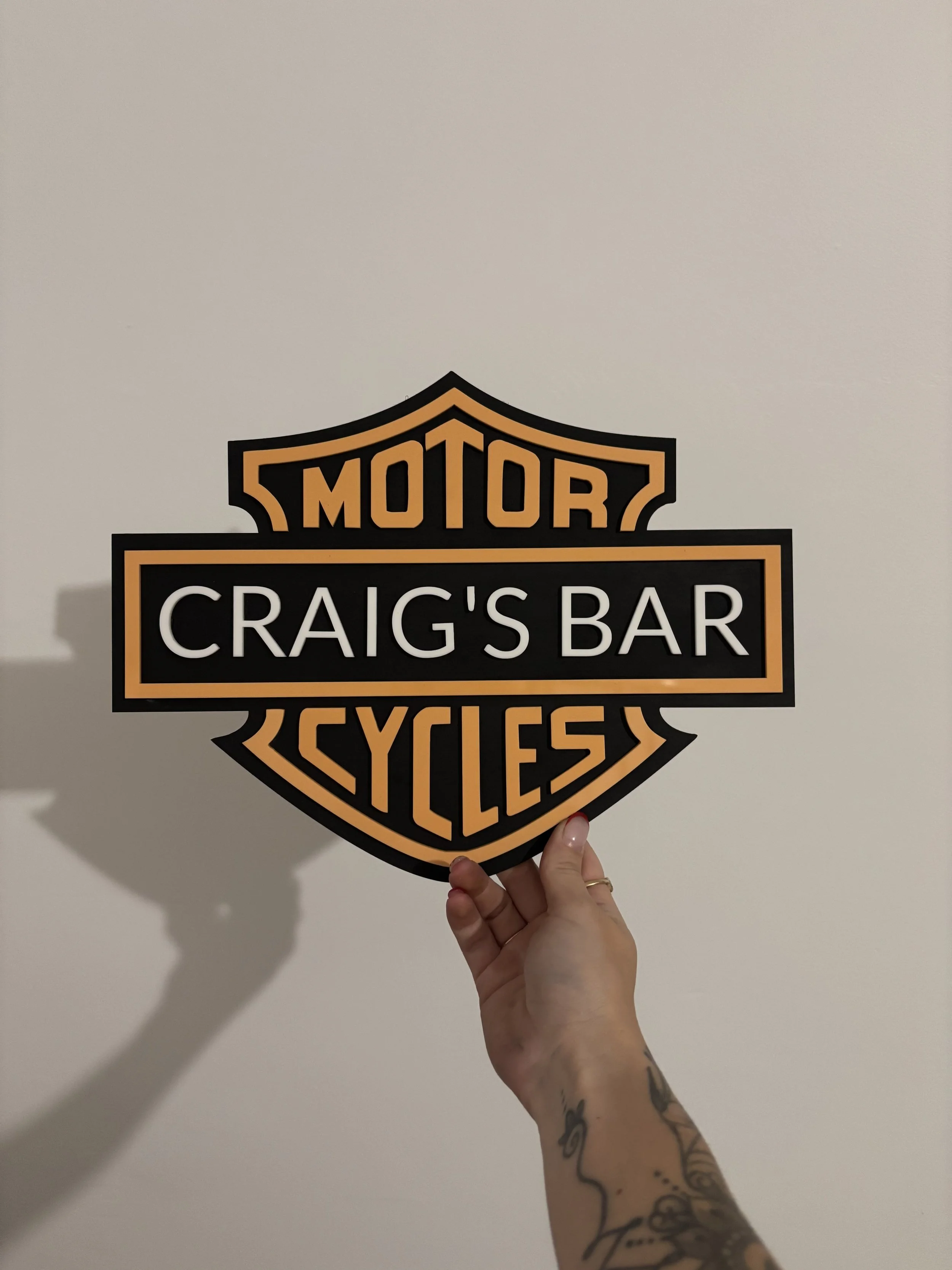 A person's hand with tattoos holding a Harley-Davidson sign that reads 'Motor Craig's Bar Bikes' against a plain white wall.