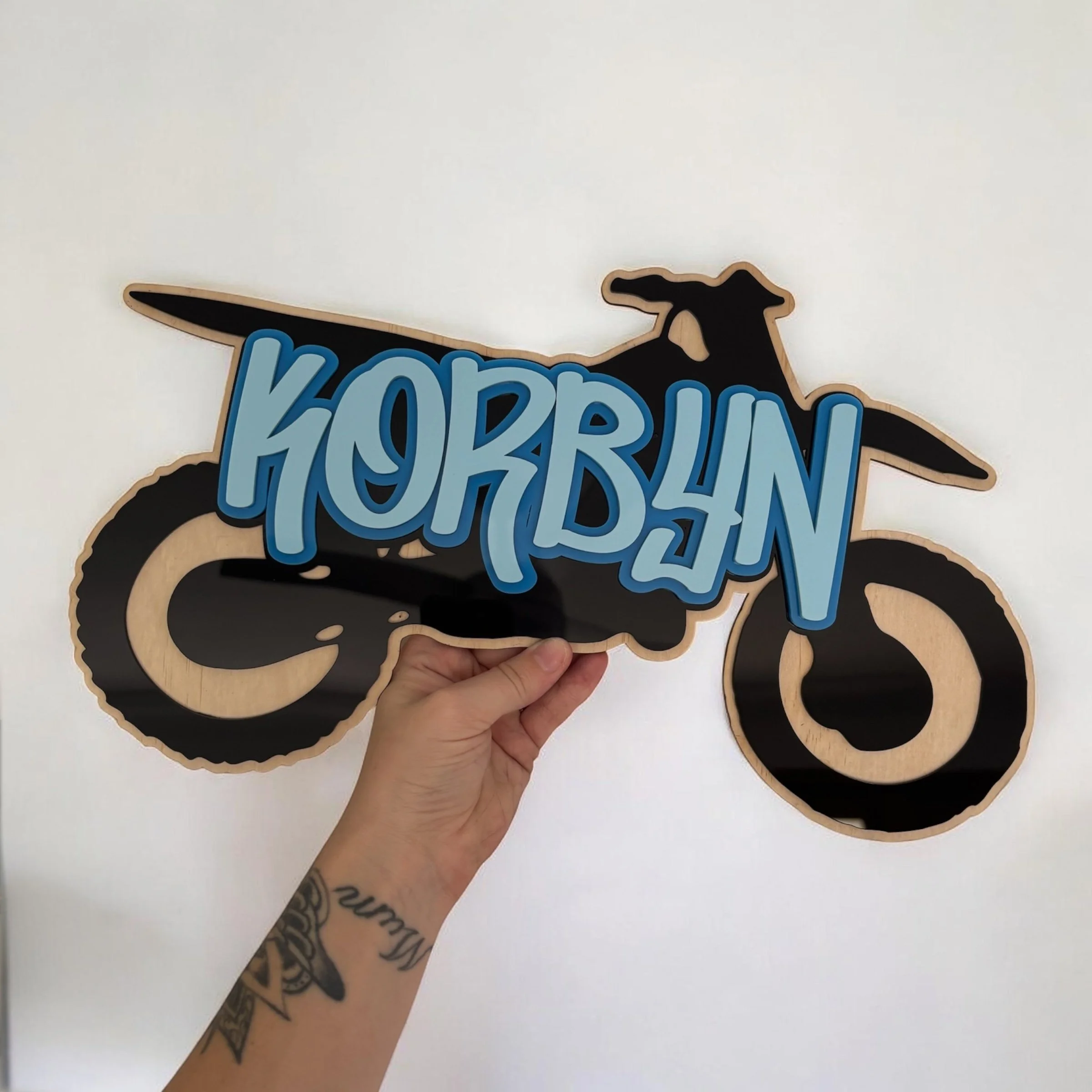 A hand holding a wooden sign with a black silhouette of a dog and blue text that reads 'HOUND' against a plain white background.