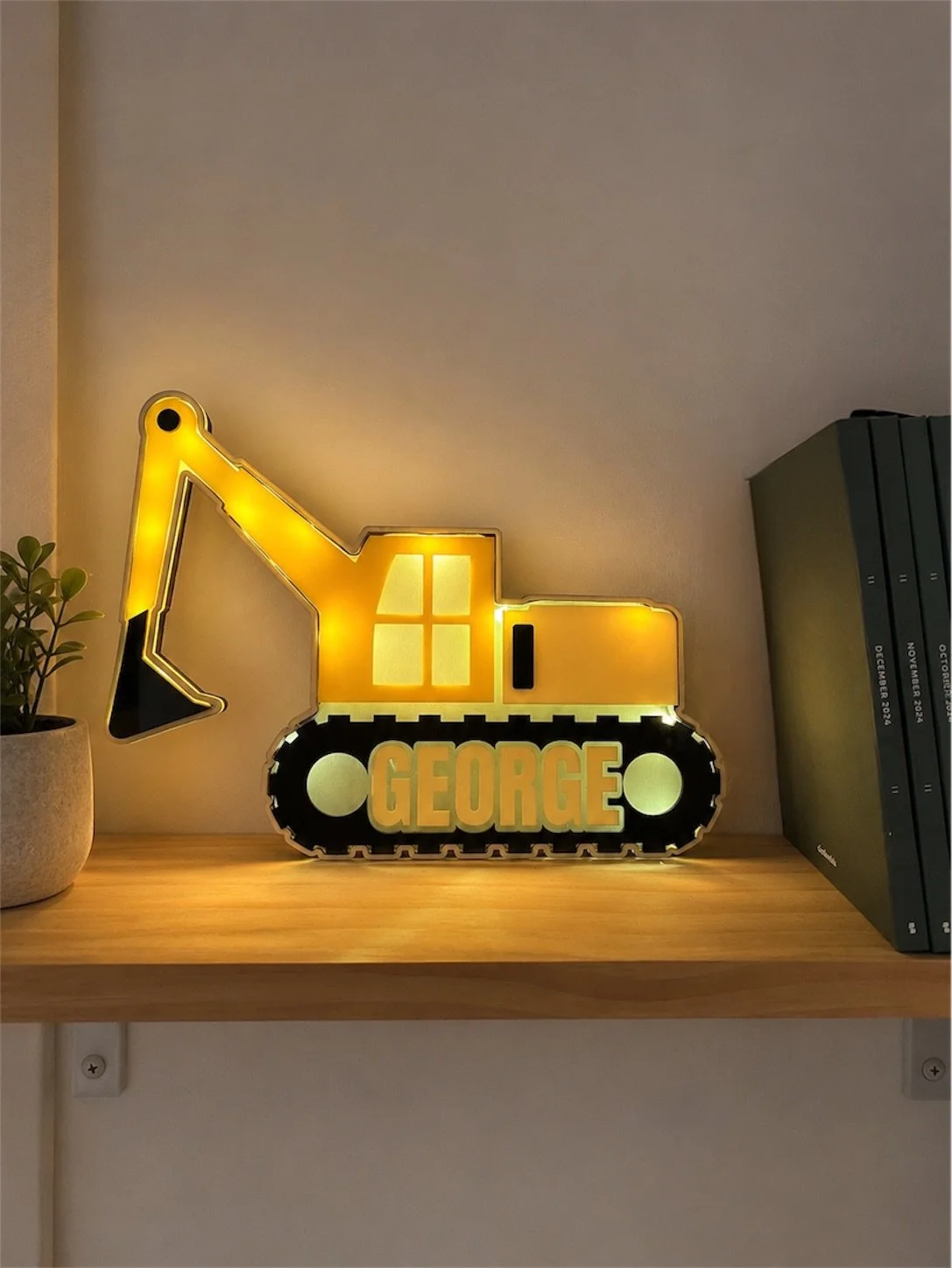 Illuminated decorative sign of a yellow excavator with the name 'George' on the tracks, placed on a wooden shelf with a potted plant on the left and a row of dark binders on the right.