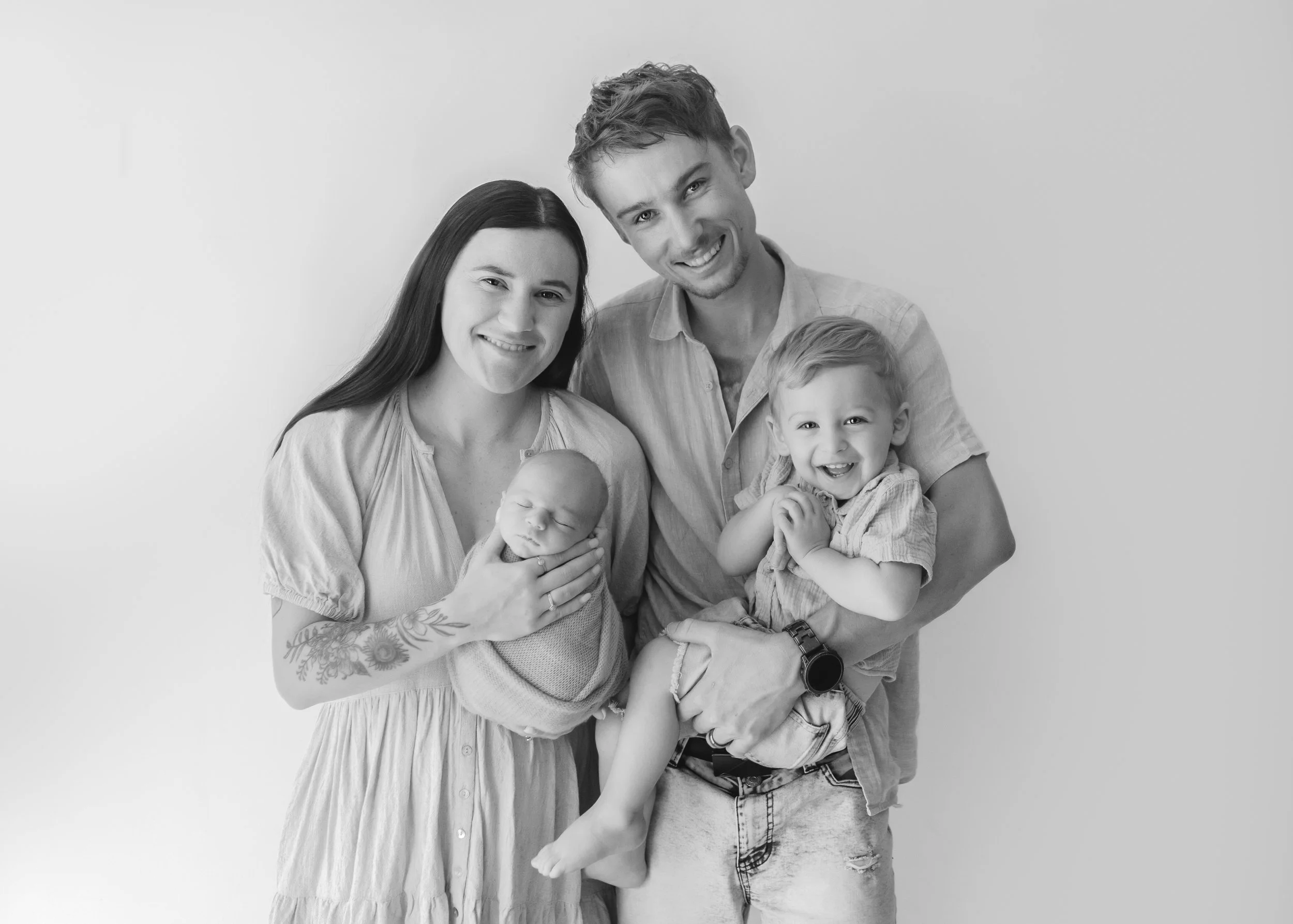A black and white photo of a smiling family of five standing together against a plain light background. The family includes a woman, a man, two young children, and an infant. The woman has dark hair and the man has light hair. The woman is holding the infant, and the young boy is being held by the man. They are dressed casually and appear happy.