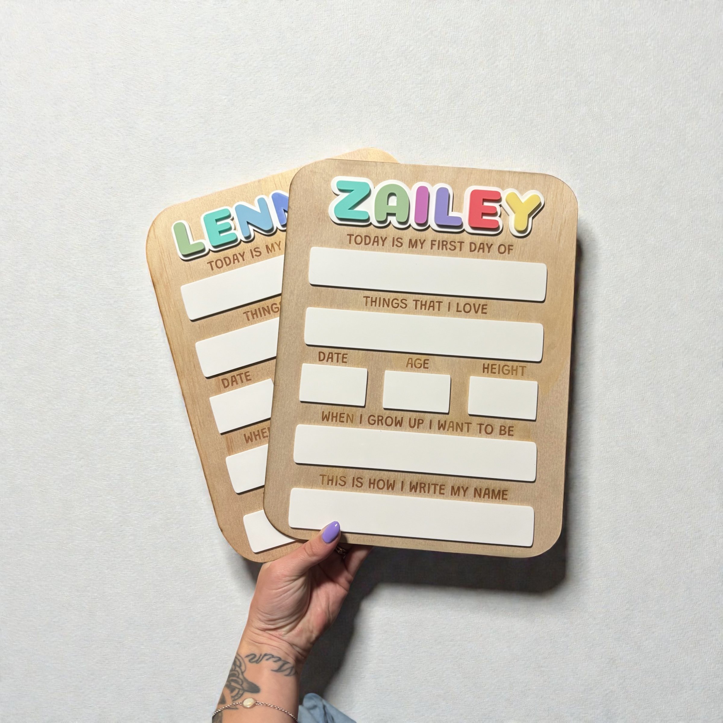Two wooden name tags with colorful lettering that spell 'LENA' and 'ZAILY,' each with a white space below for writing additional information. A person with a tattooed hand is holding the tags against a light gray background.