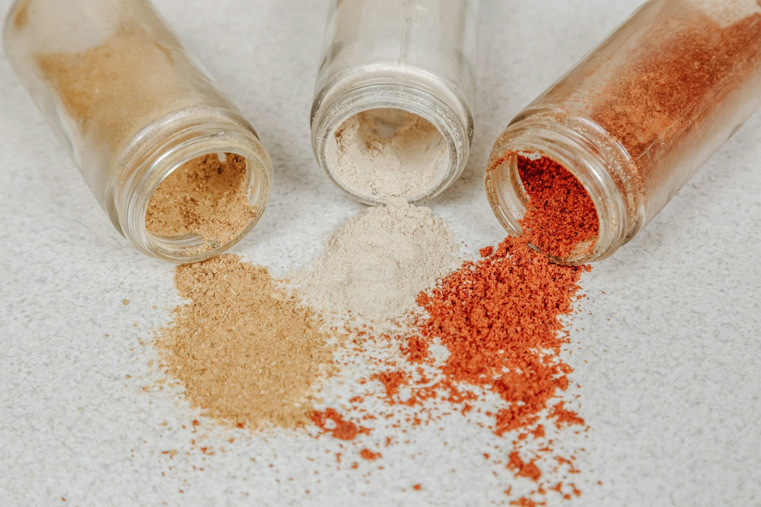 Three glass jars with open mouths lying on their sides, spilling different ground spices onto a white surface. The left jar contains a light brown spice, the middle a white or beige spice, and the right a reddish-brown spice.