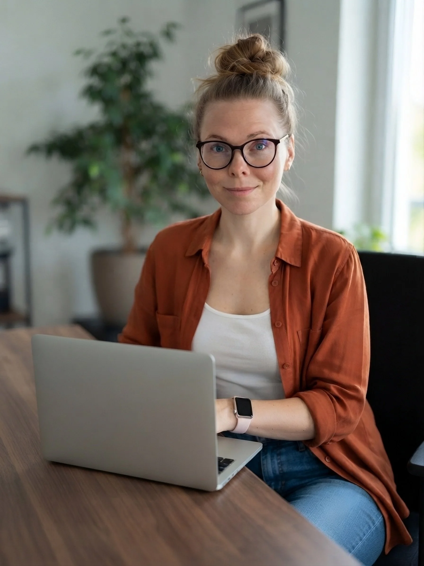 A woman with glasses and a bun hairstyle sitting at a wooden table, working on a silver laptop. She is wearing a white shirt and an orange-brown shirt, and a smartwatch on her left wrist. The background features a large houseplant, a framed picture, and a window with curtains.