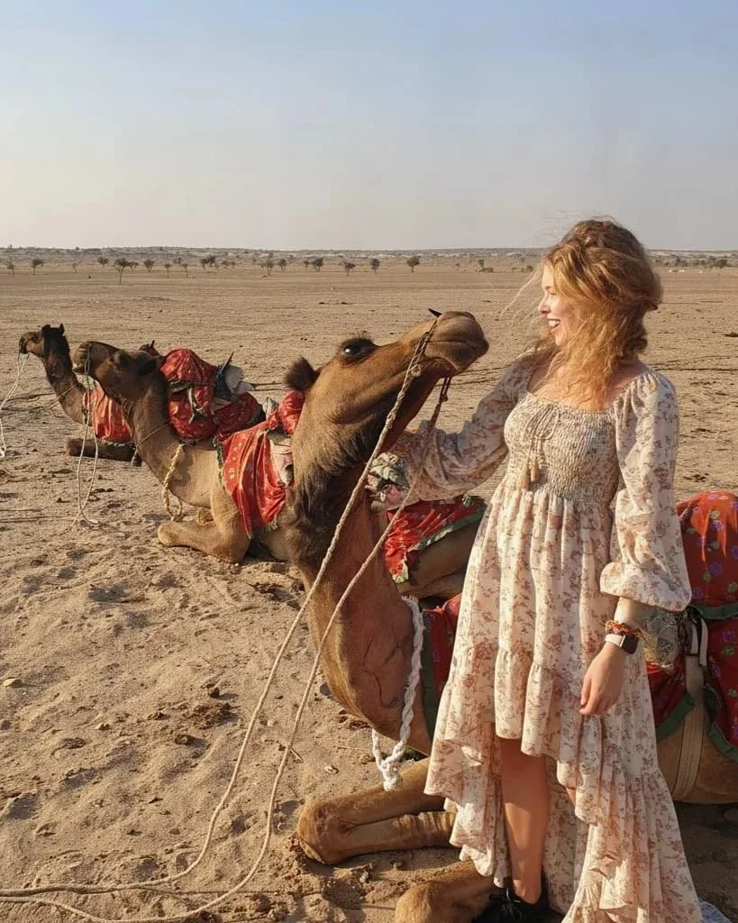 A woman in a long, floral dress smiling and holding a camel's bridle in a desert landscape with other camels sitting and adorned with colorful saddles.