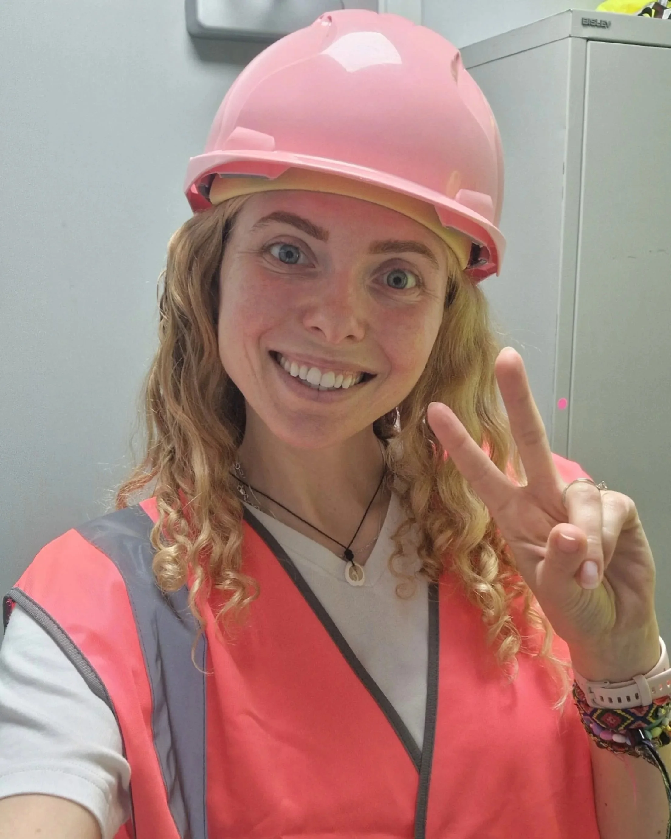 A smiling woman with curly red hair wearing a pink hard hat and a pink safety vest, making a peace sign with her right hand in an indoor setting.