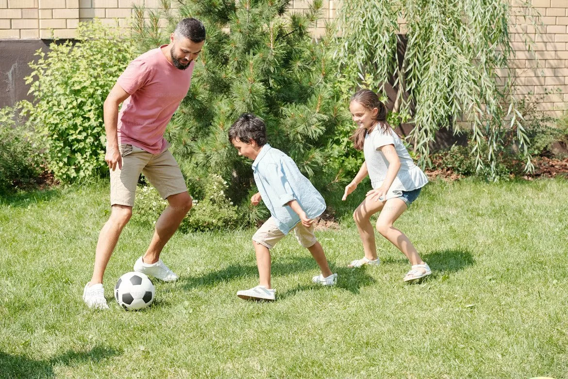 Father and two children playing soccer outdoors on a grassy yard with trees and bushes in the background.