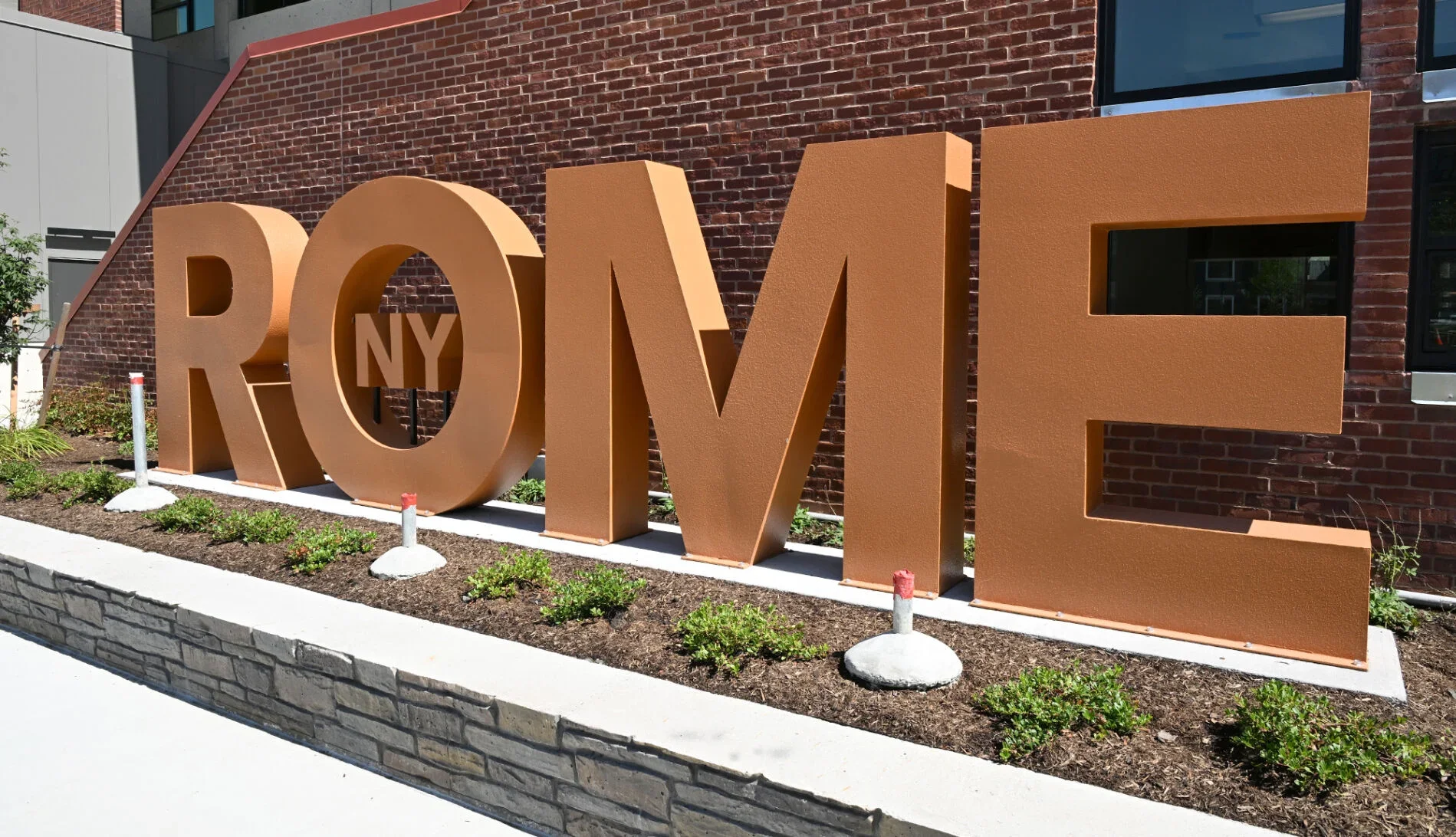 Large, three-dimensional orange sign spelling 'ROME' with a small 'NY' inside the letter 'O', placed in front of a brick building with windows and a landscaped area with plants and small white and red barriers.