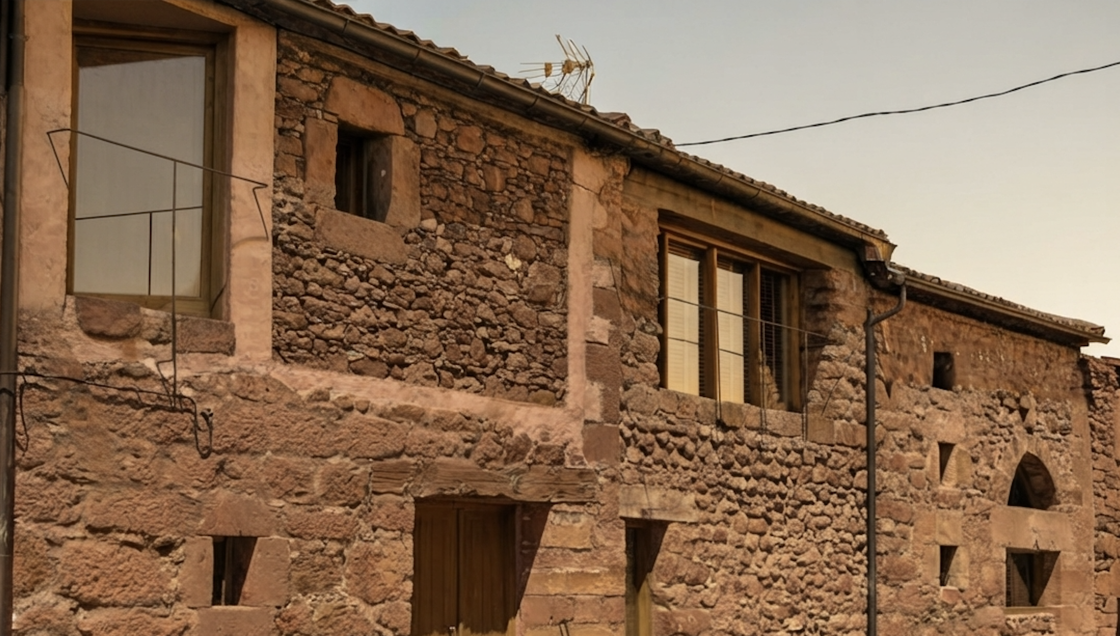 Casa rural antigua de piedra y adobe, con ventanas de madera, techo de tejas y cables eléctricos.