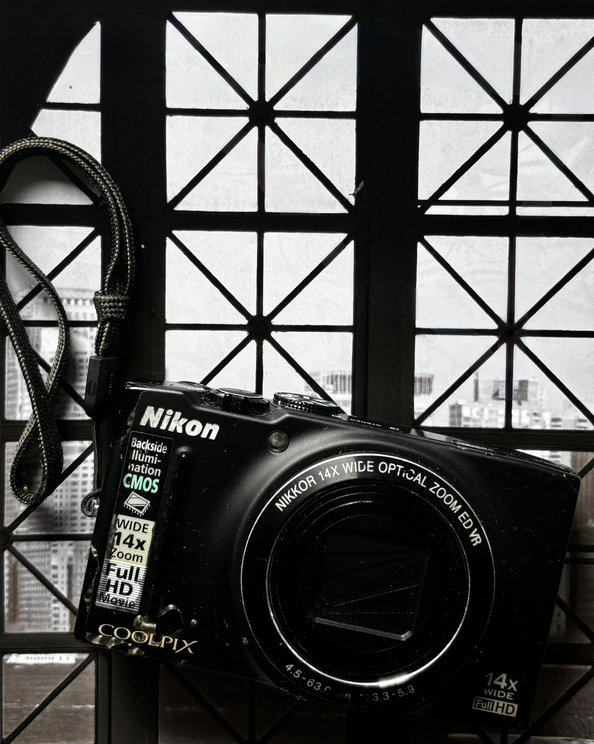 A Nikon digital camera with a black body and zoom lens, placed in front of a patterned black wire mesh window or divider. The camera's labels and specifications are visible, indicating a 14x wide optical zoom and full HD video capability. The background shows a blurred cityscape view through the window.