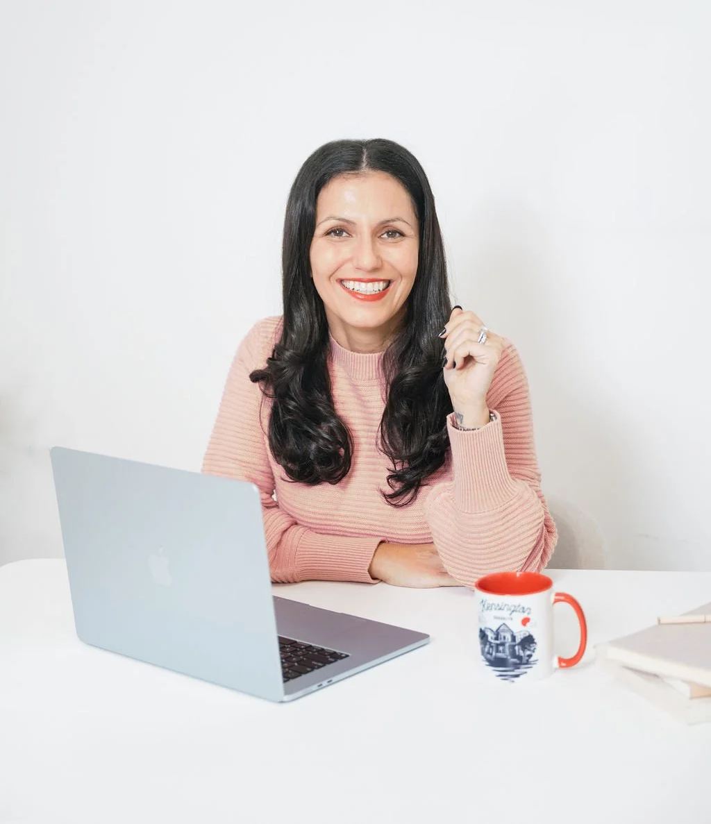 A woman with long dark hair smiling while sitting at a white desk with a laptop, a red and white mug, and some notebooks.
