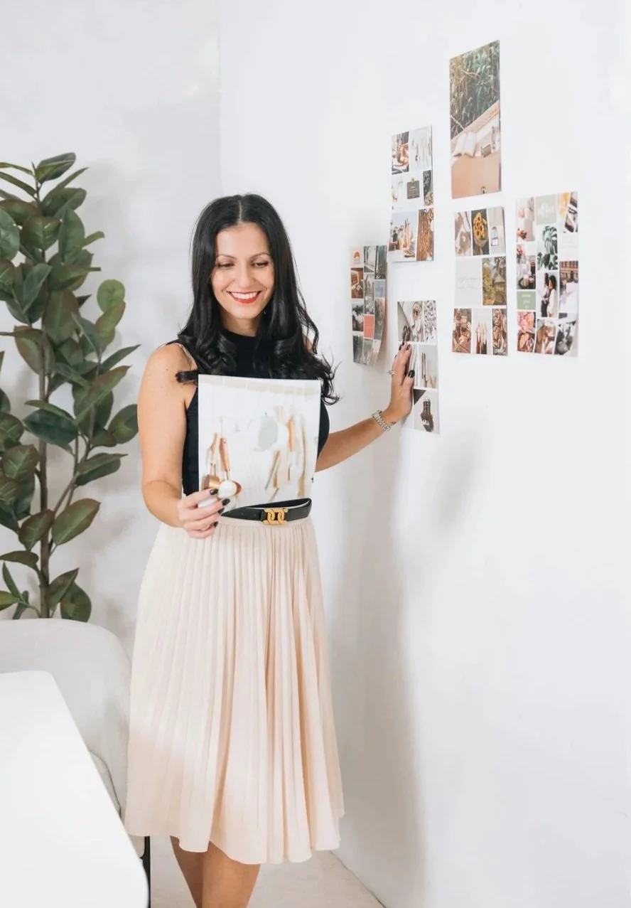 Smiling woman in a black top and pleated beige skirt holding a photo and standing near a white wall decorated with small photos. A large green plant is on her left.