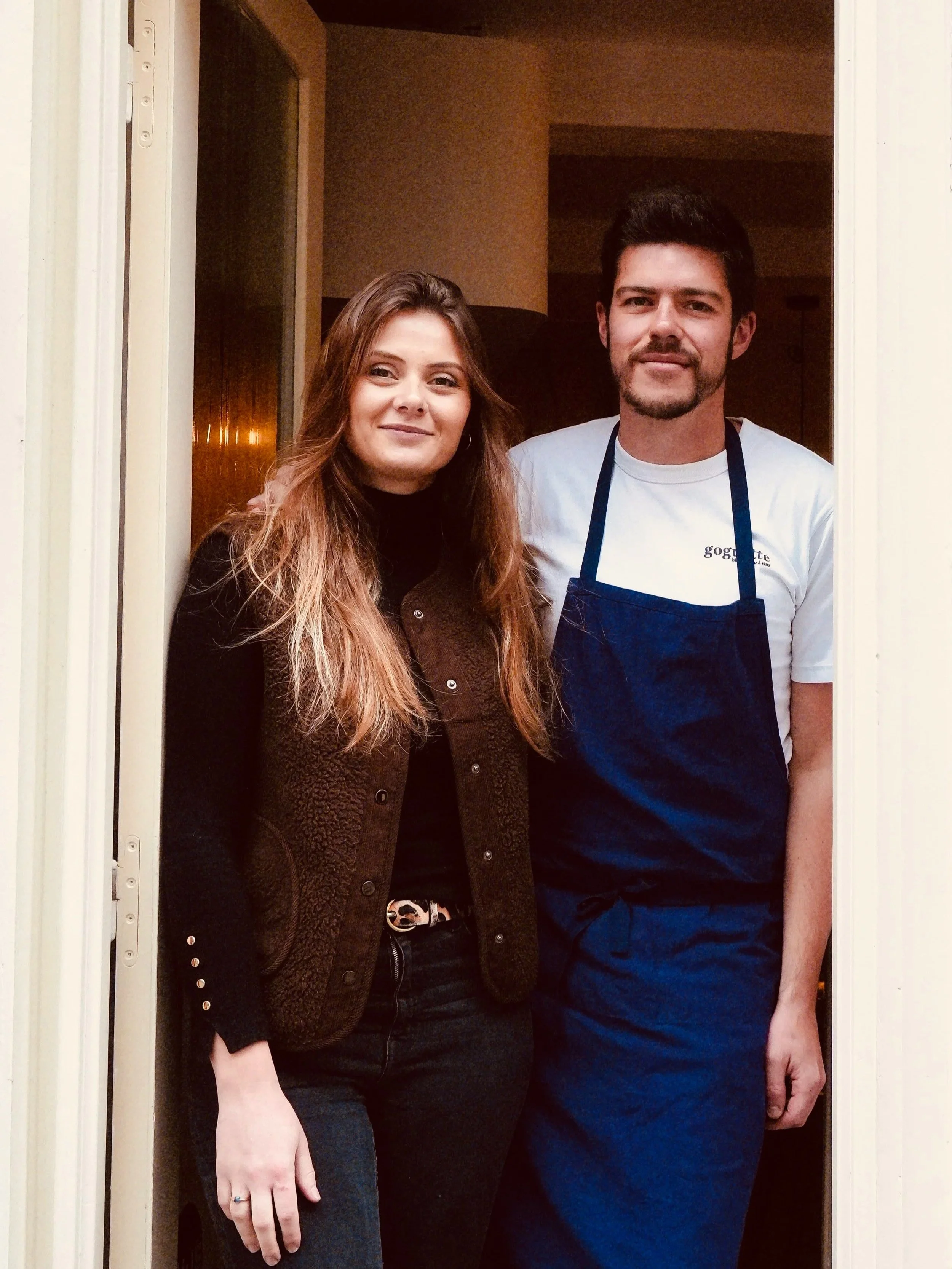 Deux personnes, un homme et une femme, posent dans une porte. La femme a de longs cheveux bruns et porte une veste marron, tandis que l'homme a des cheveux noirs, une barbe et porte un t-shirt blanc avec un tablier bleu.