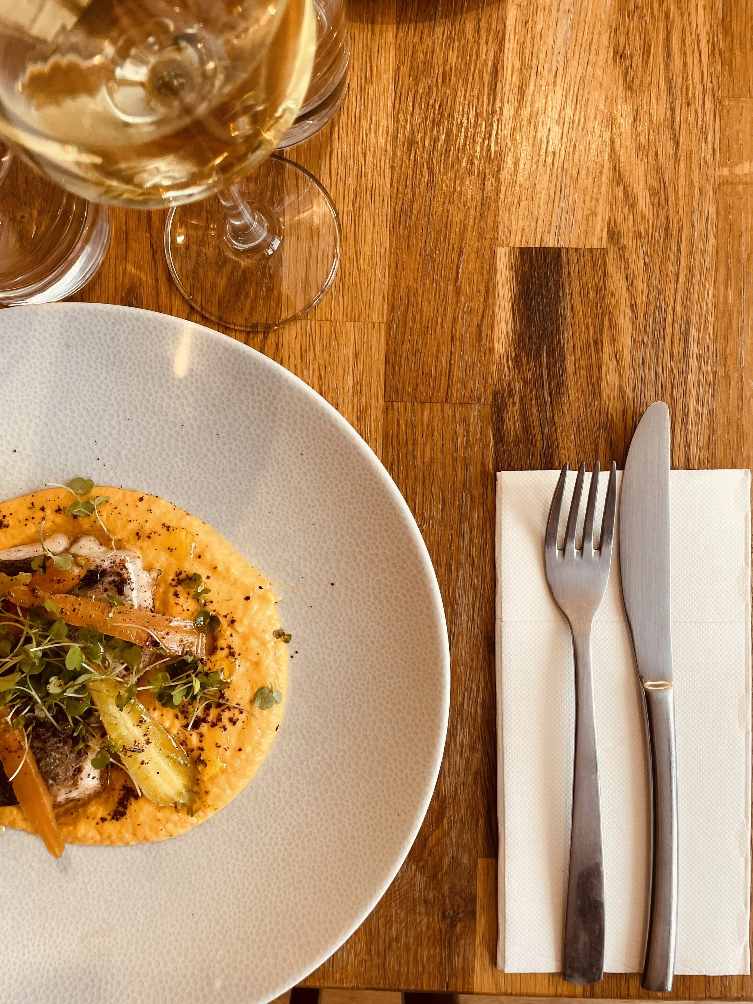 Assiette de nourriture avec une sauce orange, microgreens, et des légumes.
Verres de vin blanc et de l'eau sur une table en bois, avec un couteau, une fourchette, et une serviette blanche.