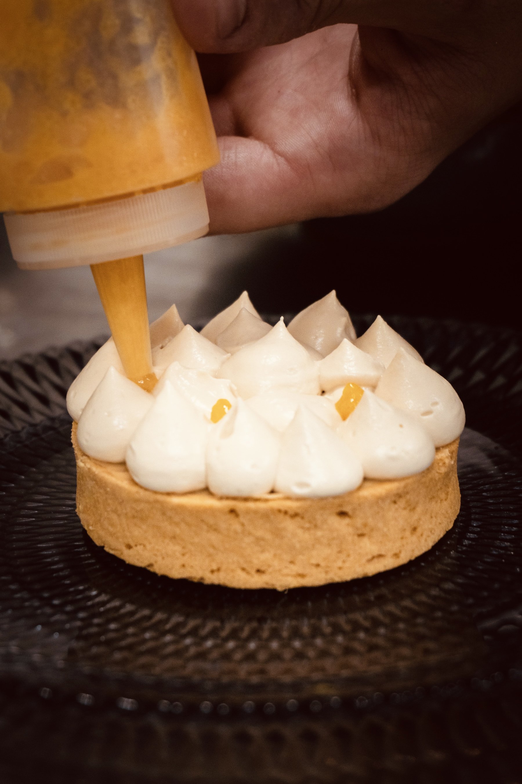 Une personne verse de la crème ou du glaçage dans un gâteau rond à la cerise tart. Le gâteau est placé sur une surface sombre et comporte des décorations de petites touches de coulis ou de confiture.