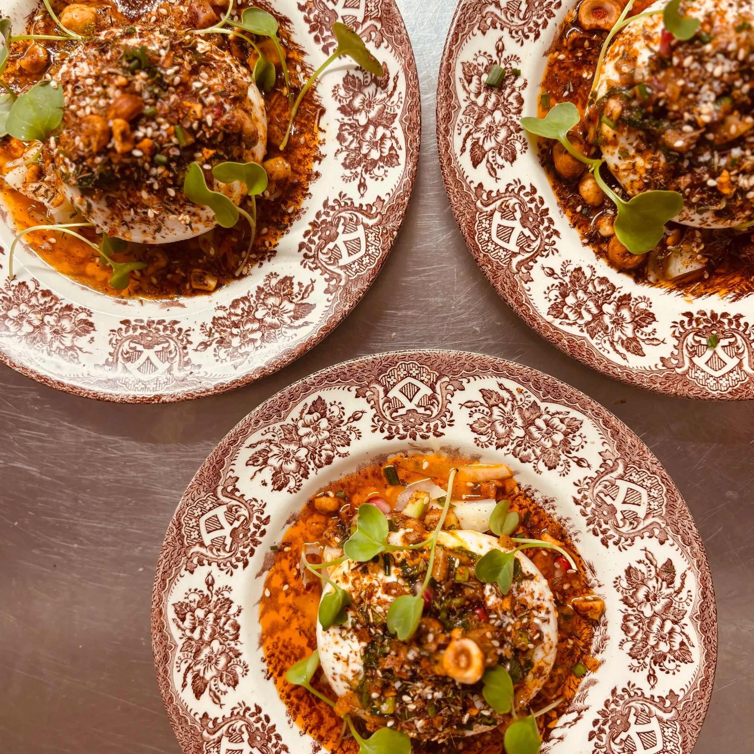 Œufs mollets recouverts d'une sauce épicée avec graines de sésame, herbes et micro-pousses, présentés sur une assiette décorative.