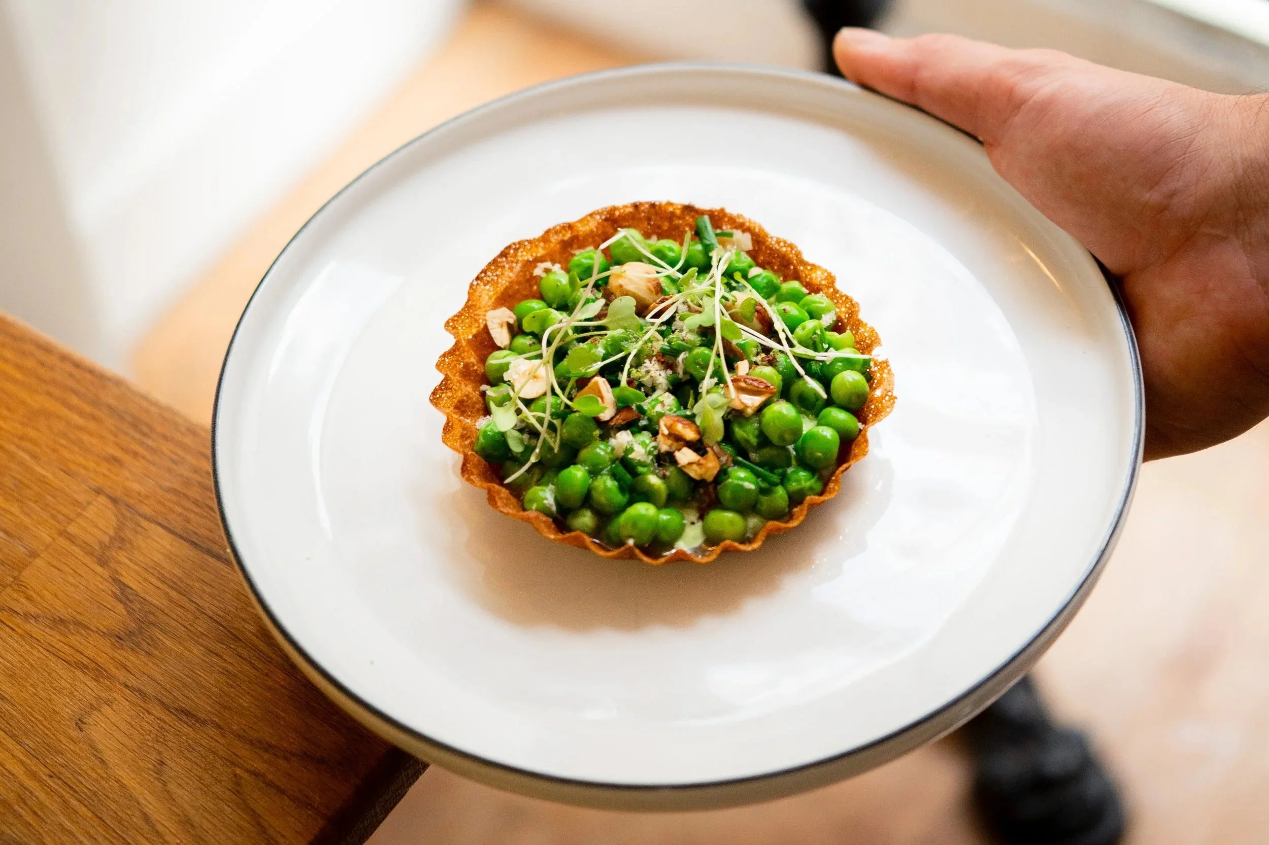 Une tartelette croustillante garnie de petits pois, herbes, noix et fromage râpé, servie sur une assiette blanche. Une main tient l'assiette, posée sur une surface en bois.