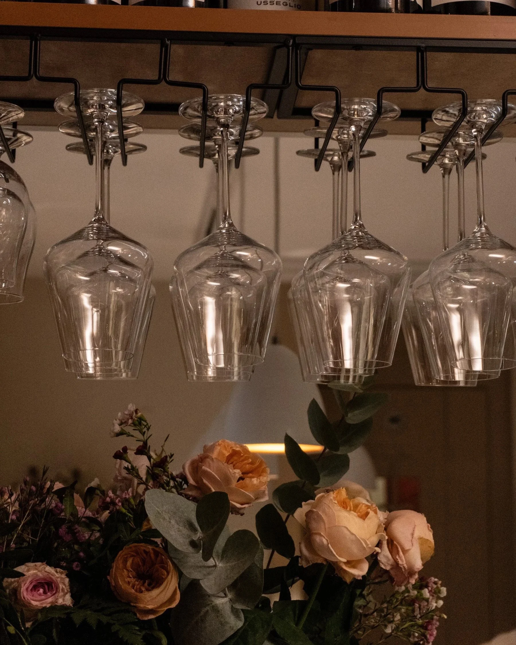Vase suspendu avec plusieurs verres à vin à l’envers sous une étagère en bois. En dessous, un arrangement floral avec des roses, des feuillages et des fleurs roses et blanches, dans un décor intérieur chaleureux.