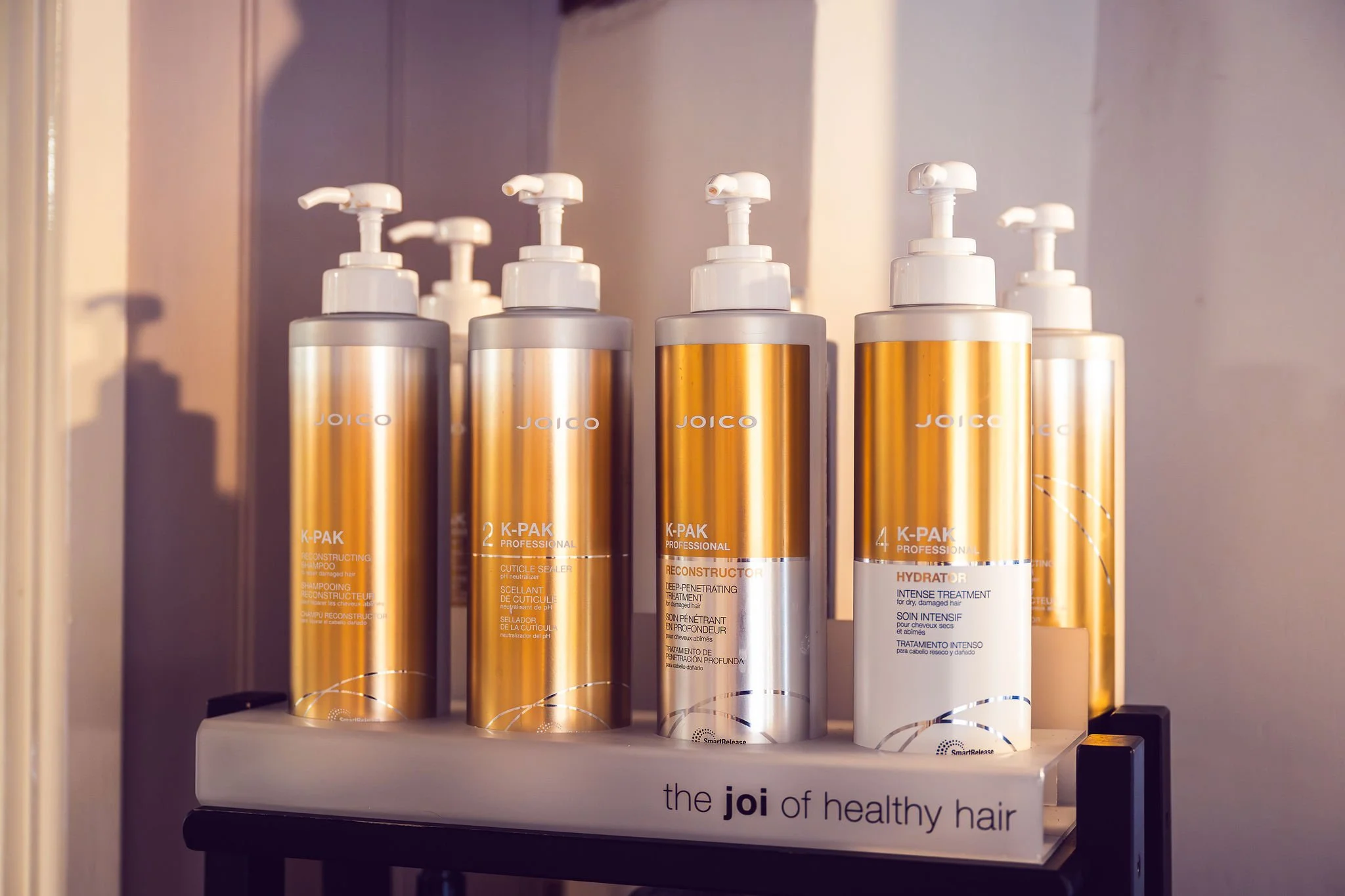 Several bottles of Joico hair care products, including reconstructors and hydrators, are arranged on a shelf with a label that reads 'the joi of healthy hair'.