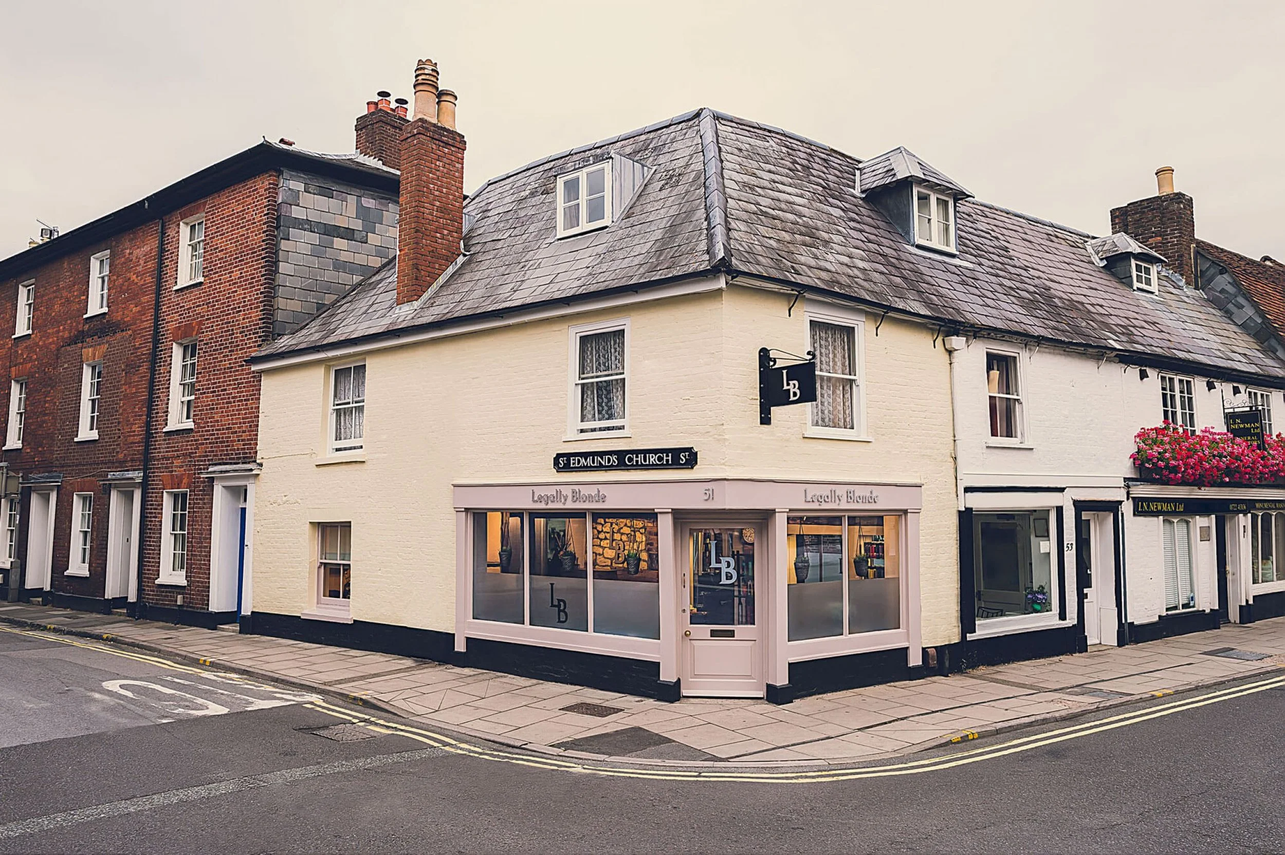 Street view of a white building with a storefront named 'Legally Blonde' on the ground floor, above which there are residential windows and a slate roof with dormer windows and chimneys. Next to it, there's a brick building with multiple windows and a chimney.