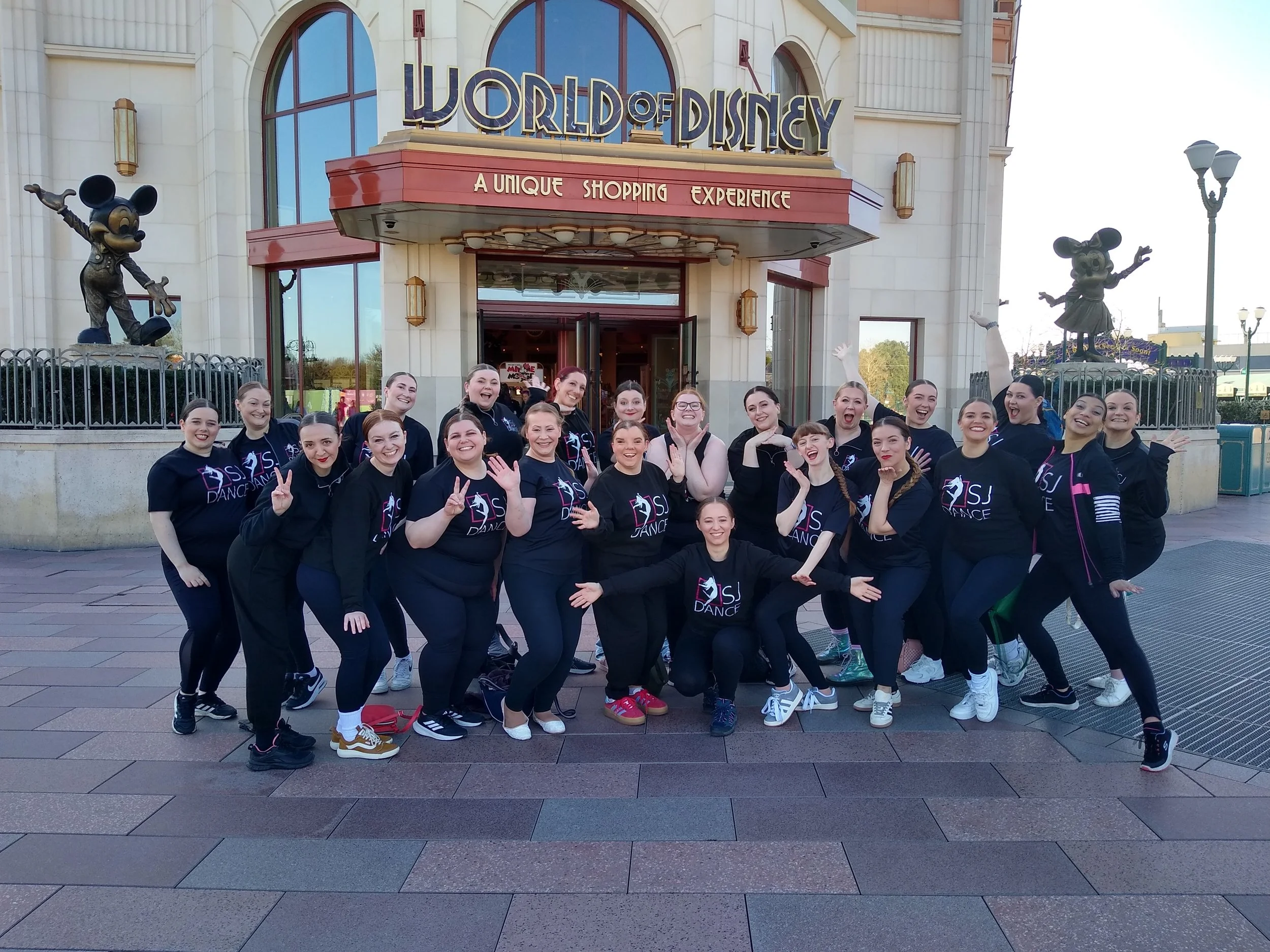Group of dancers in black T-shirts with a logo, smiling and posing in front of Disney's World of Disney store. The store has large glass windows, an arched entrance, and statues of Mickey Mouse characters.