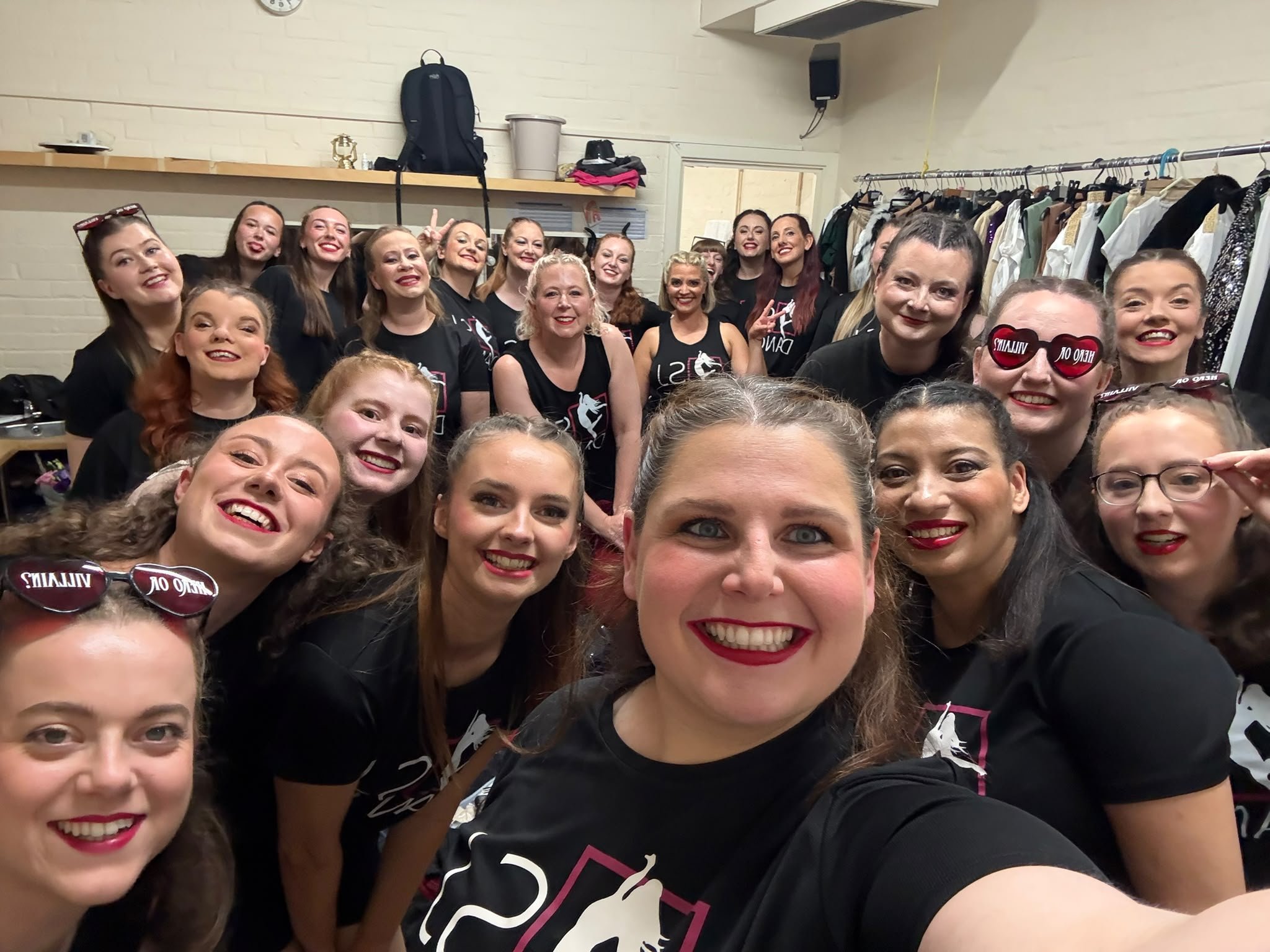 A large group of women wearing black shirts, smiling for a selfie in a dressing room with clothes on racks and a high ceiling.