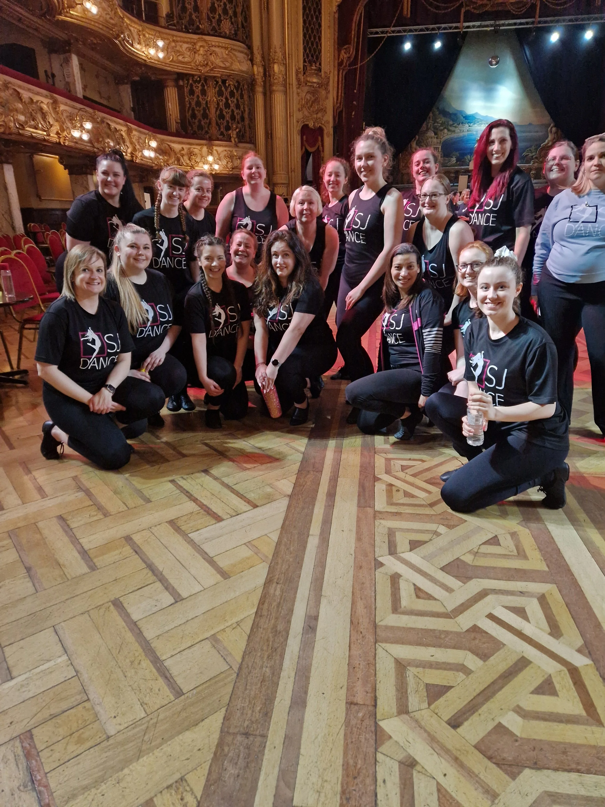 A group of women in black and gray dance shirts are posing together on a stage inside a grand theater with ornate gold decorations, wooden flooring, and a painted backdrop of a river landscape.