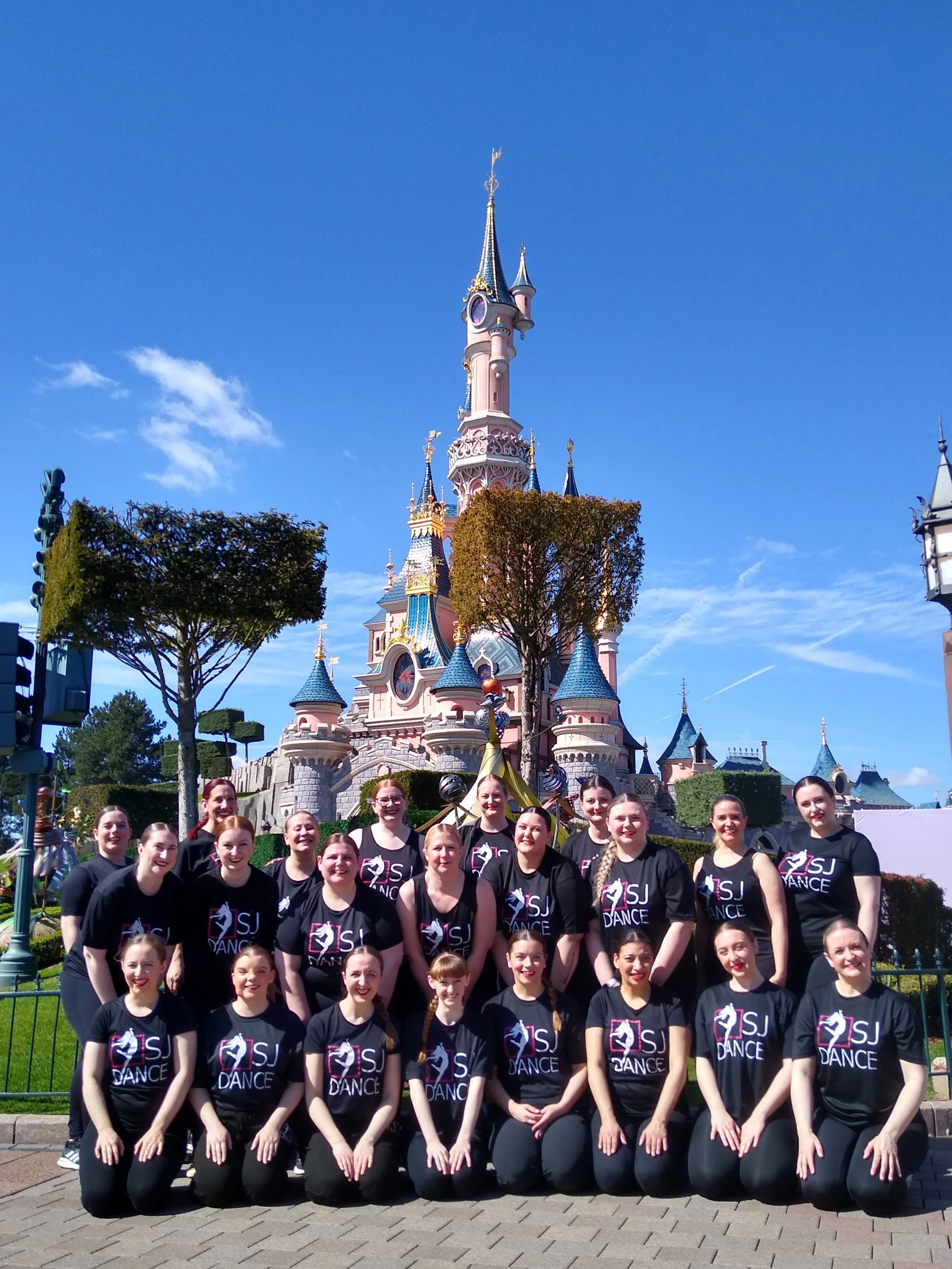 A group of young female dancers in black shirts with the logo 'SJ Dance' are posing in front of a castle at Disneyland. The castle has pink and blue spires and is set against a bright blue sky with a few clouds.