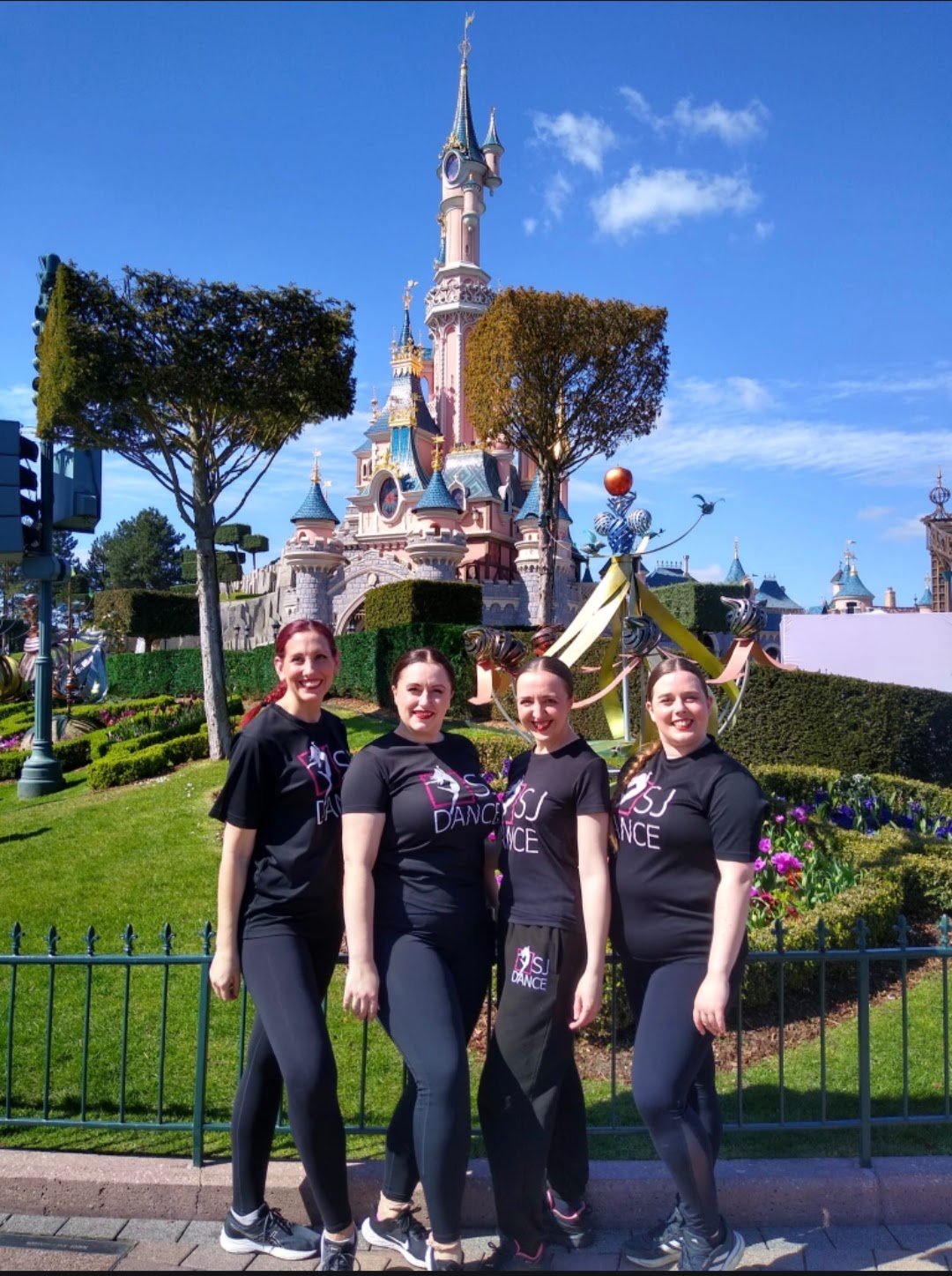 Four women, dressed in black dance shirts and black leggings, posing in front of Sleeping Beauty Castle at Disneyland with manicured gardens and a blue sky in the background