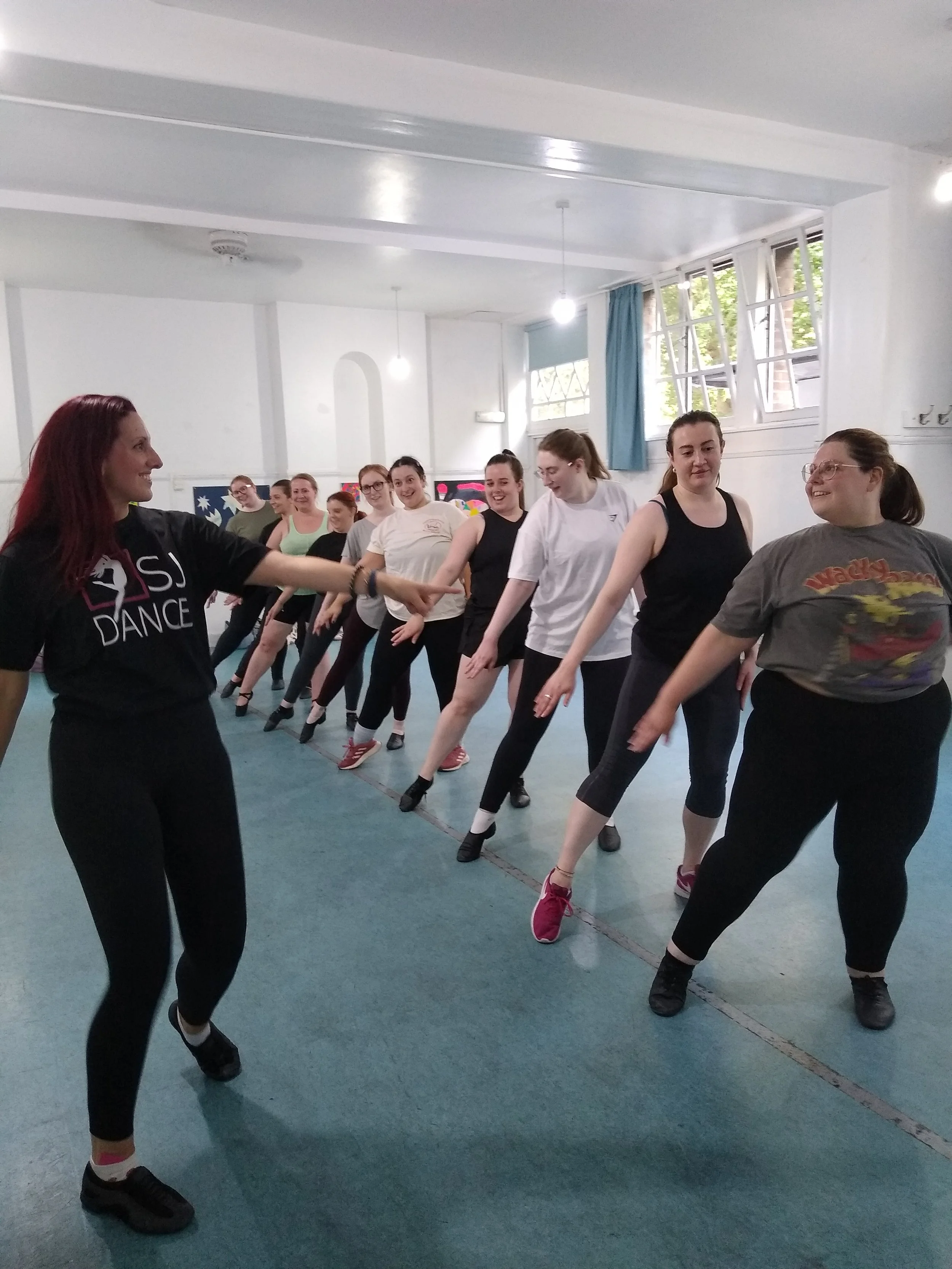 Group of women participating in a dance class in a studio, standing in a line, with a dance instructor at the front.