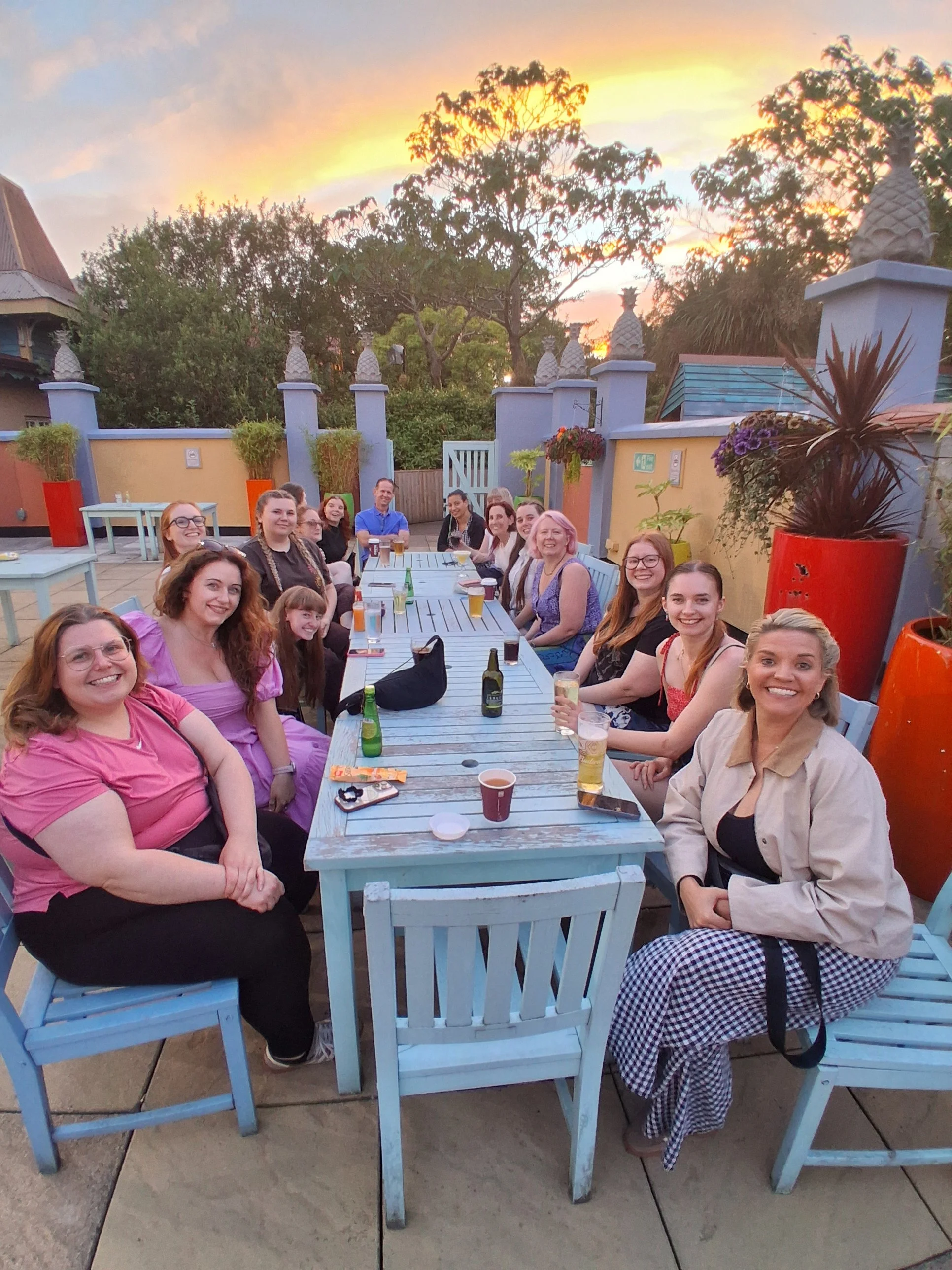 People sitting at a long outdoor table on a terrace during sunset, smiling and enjoying drinks, with colorful planters and greenery in the background.