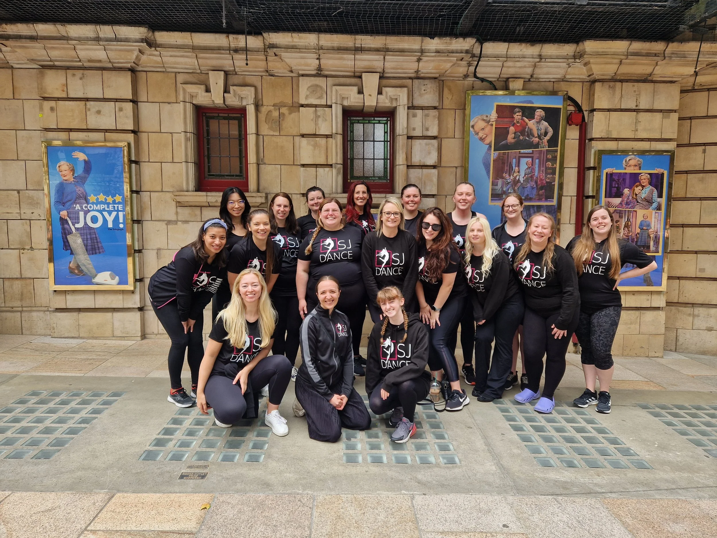A group of smiling young women wearing black T-shirts with a ballet dancer logo, standing in front of a historical building with stone walls and decorative windows. There are also framed posters with images of an older woman and other scenes mounted on the wall behind them.