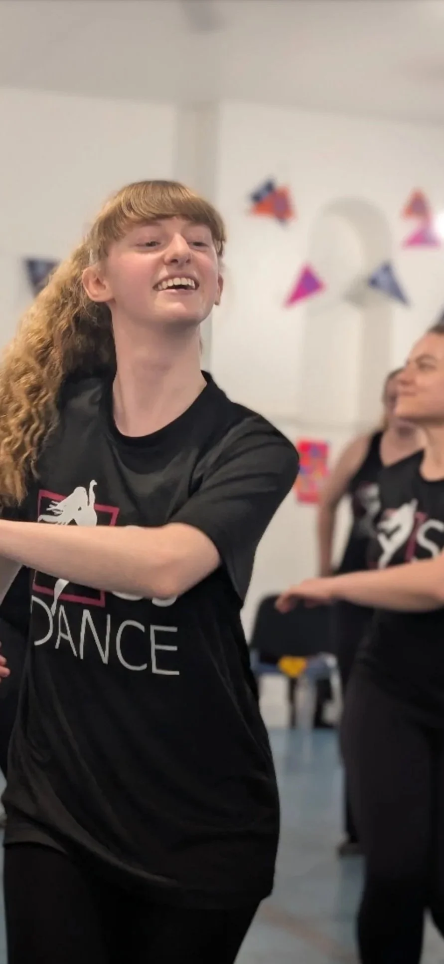 A young woman with curly red hair smiling and dancing in class, wearing a black T-shirt. In the background, another woman is visible, also in a black T-shirt, with colourful decorations hanging in the room.