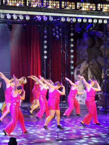 Group of women in pink costumes performing a dance on stage with red curtains and stage lights.